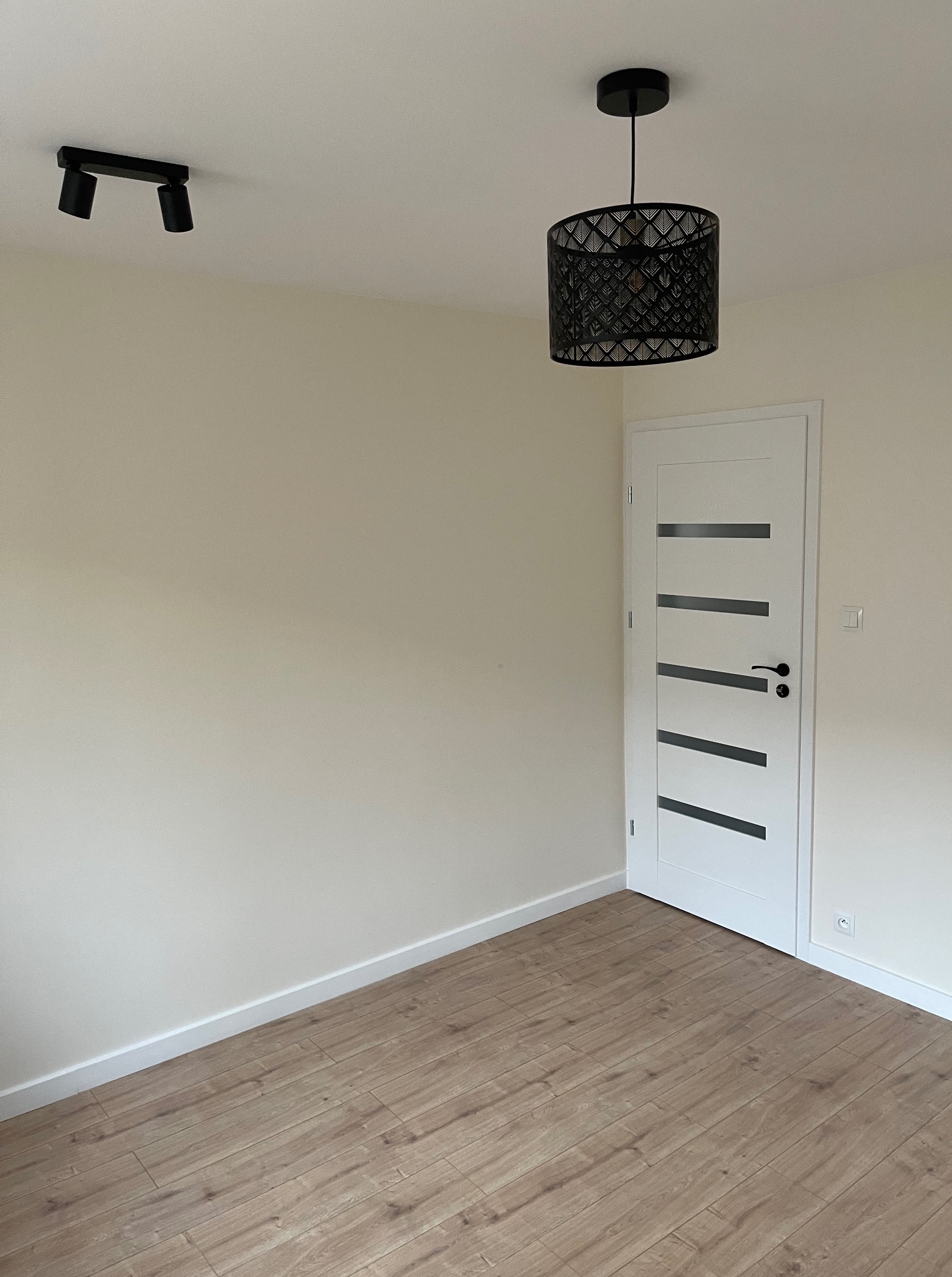 Jasne wnętrze po remoncie: panele podłogowe, kremowe ściany, białe drzwi z czarnymi akcentami, lampa sufitowa i reflektor. Minimalistyczny design.