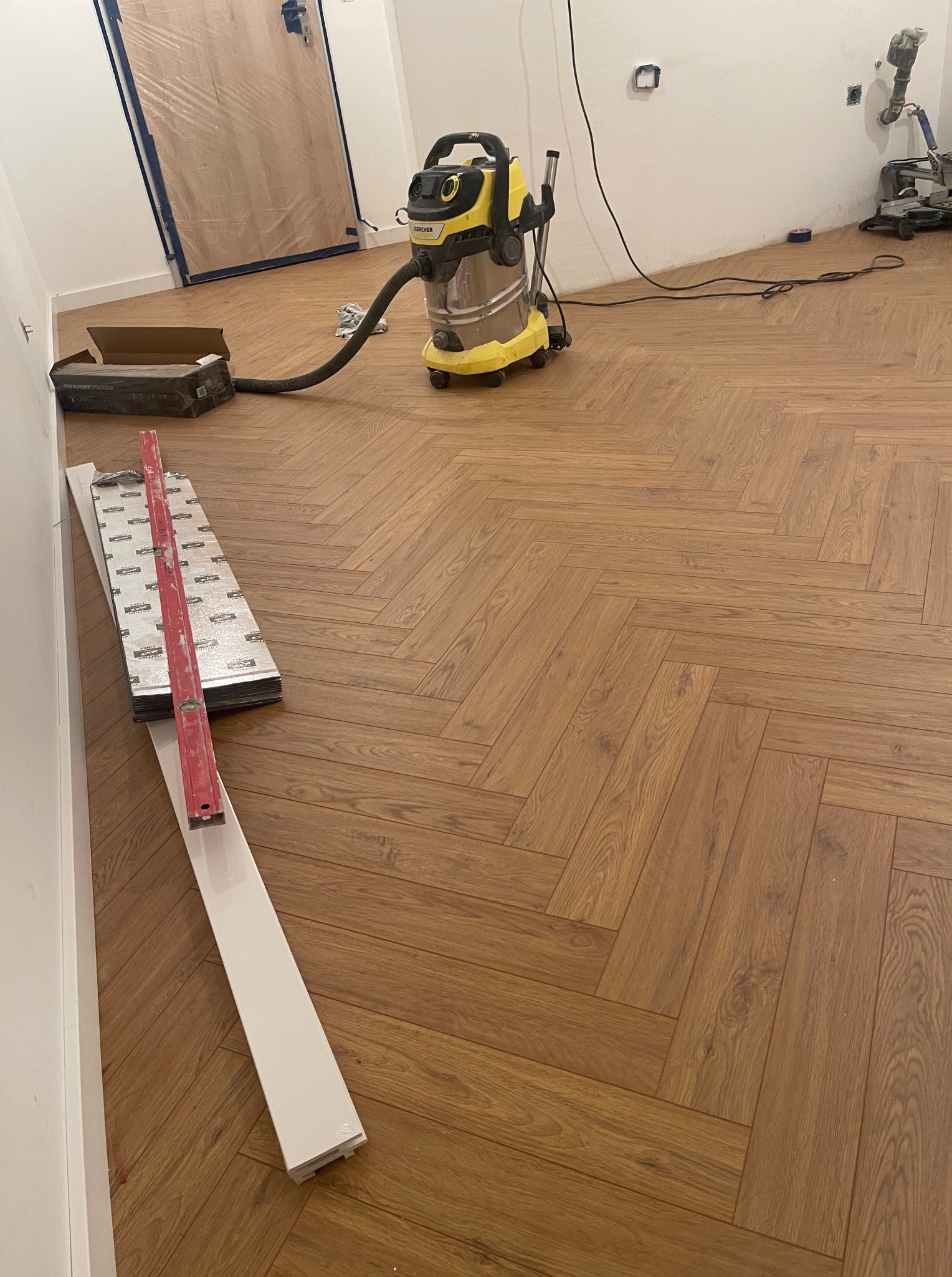 Ułożona podłoga z paneli w jodełkę, poziomica, listwa przypodłogowa, odkurzacz przemysłowy. Widok na fragment pomieszczenia w trakcie prac wykończeniowych.