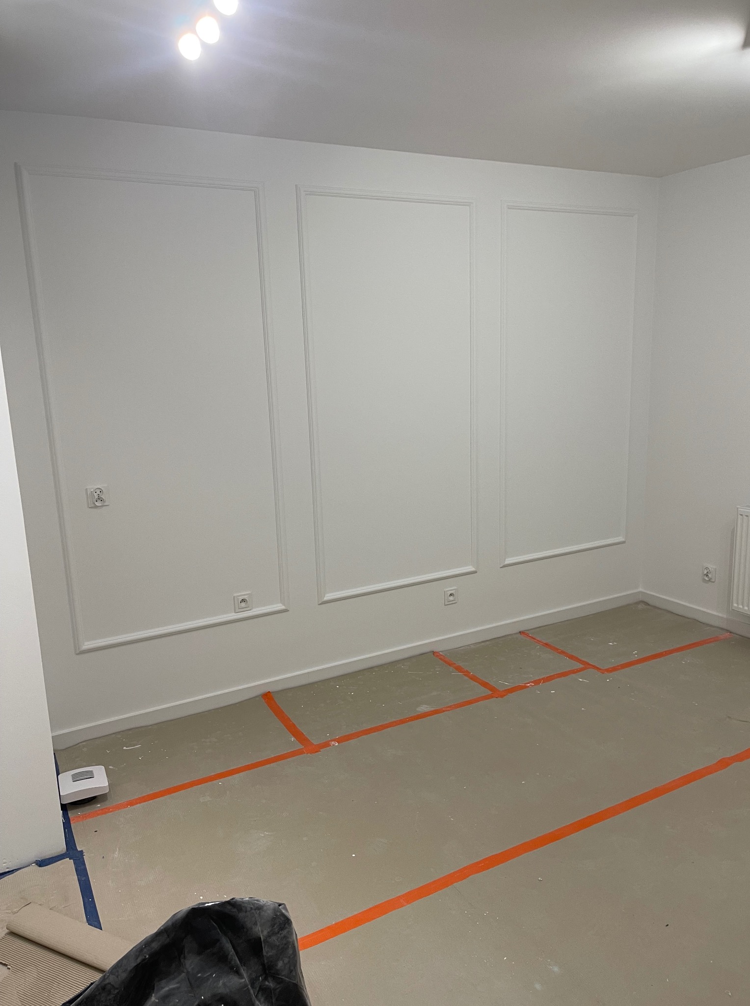 Białe ściany z dekoracyjnymi listwami w pustym pokoju, przygotowanym do układania paneli. Widoczne taśmy wyznaczające obszar roboczy na podłodze.