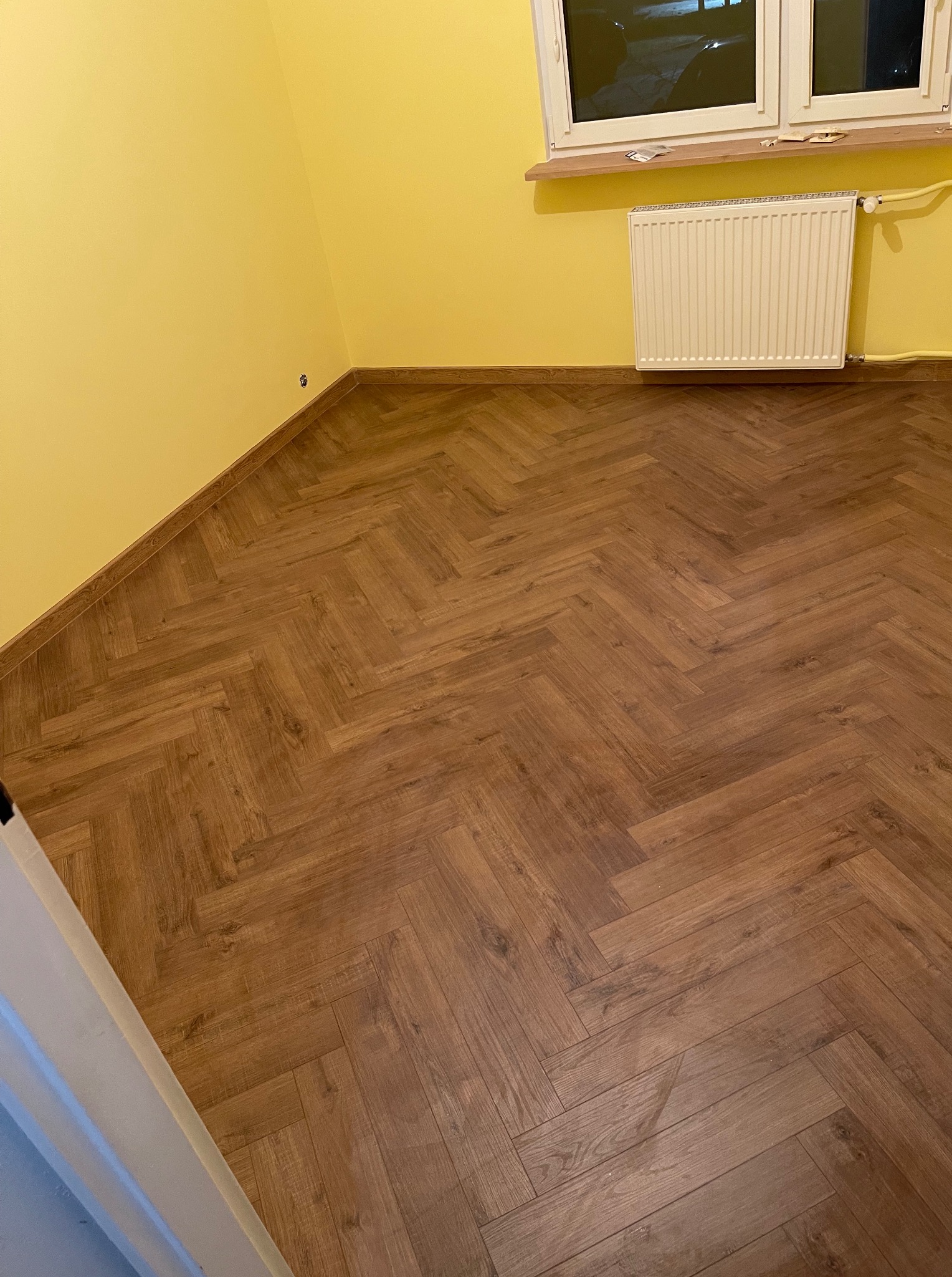 Ukończone układanie paneli w jodełkę w pokoju z żółtymi ścianami. Widoczny grzejnik i okno. Nowa podłoga z drewnopodobnych paneli. Listwy przypodłogowe zamontowane.