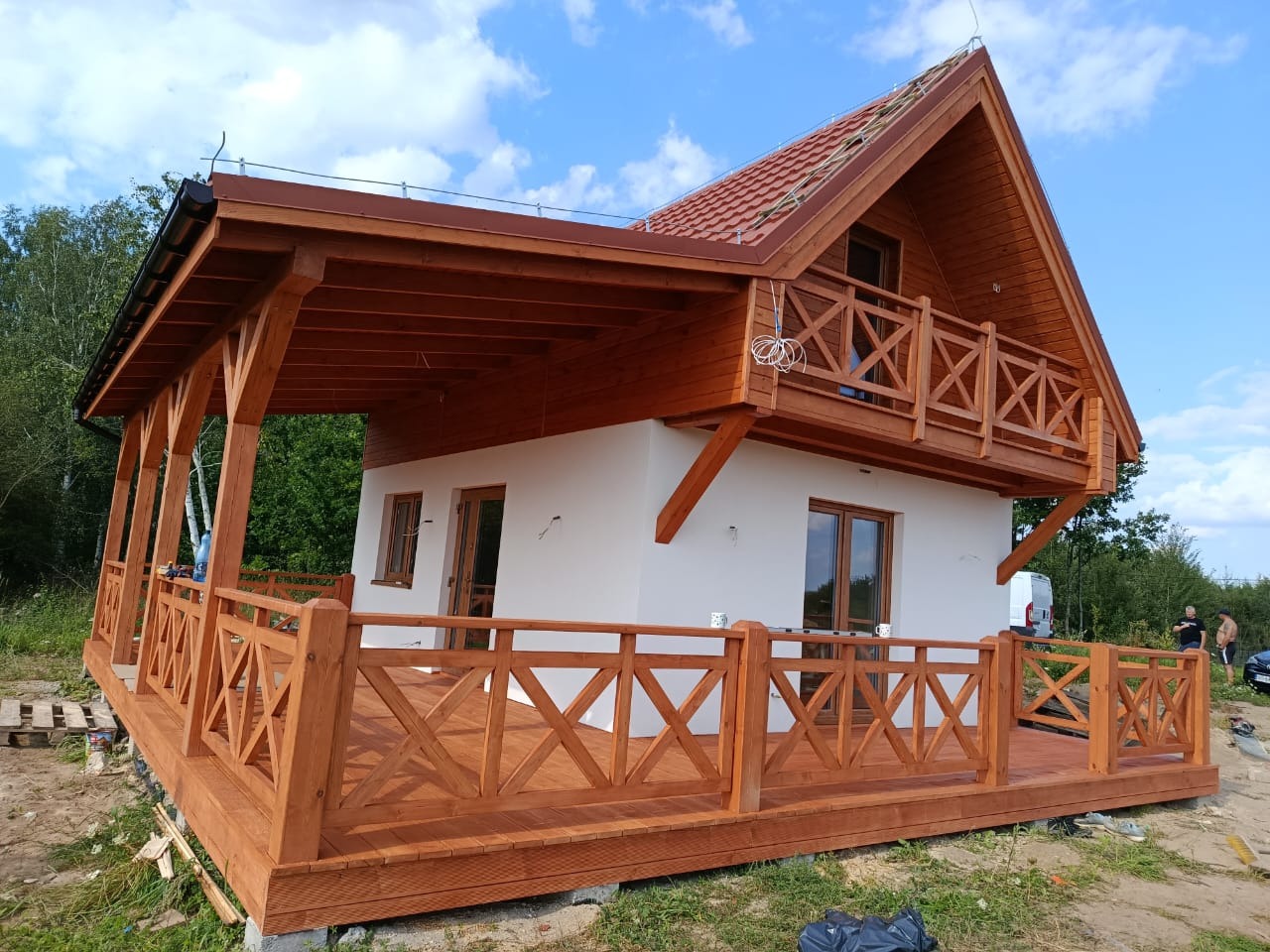 Nowoczesny dom szkieletowy z drewnianym tarasem i balkonem, wykończony w ciepłych odcieniach brązu, na tle zieleni i błękitnego nieba.