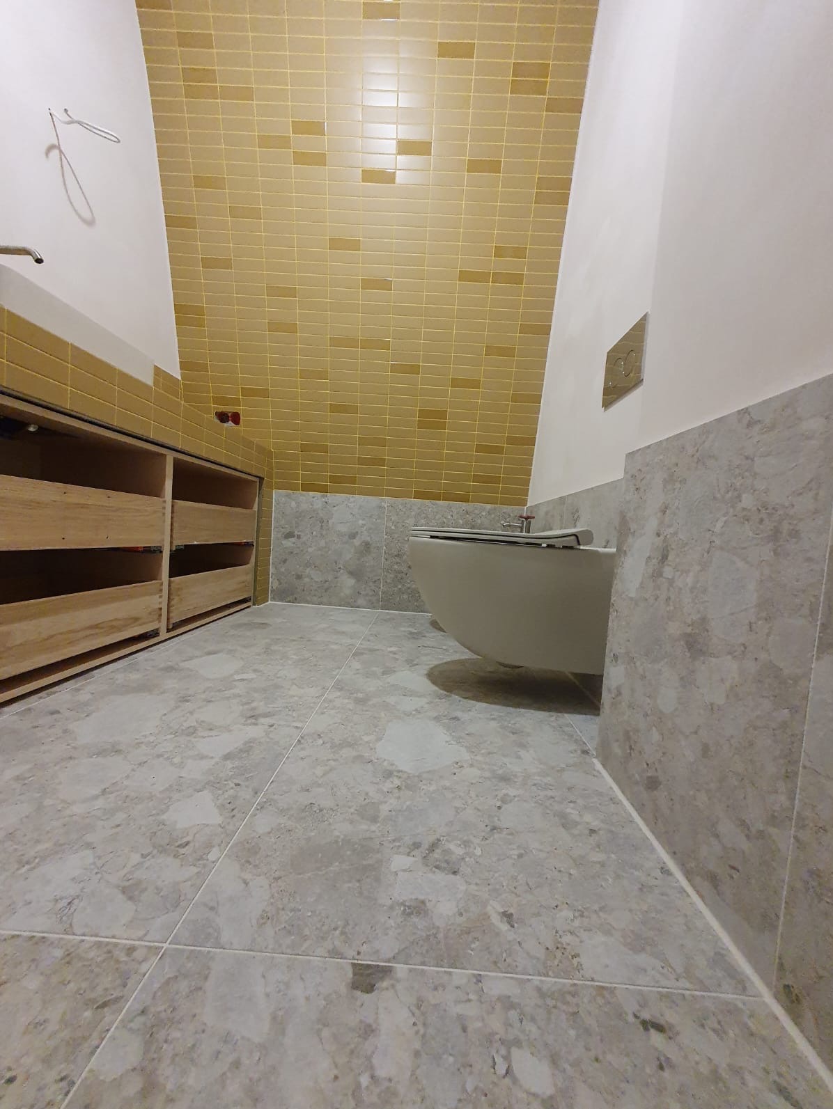 Wykończona łazienka z beżowymi płytkami na suficie i ścianie, szarą podłogą i podwieszaną toaletą. Widoczne drewniane szafki bez frontów.