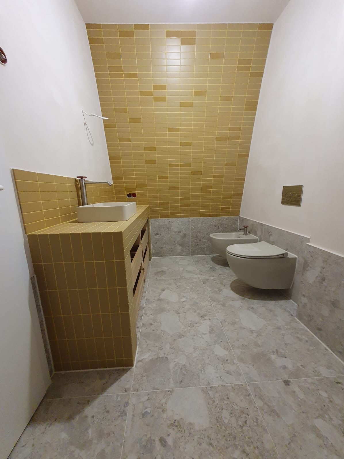 Żółta mozaika w łazience: ściana, zabudowa umywalki i blat z umywalką. Szare płytki podłogowe i fragment toalety. Ukośna ściana z mozaiką.