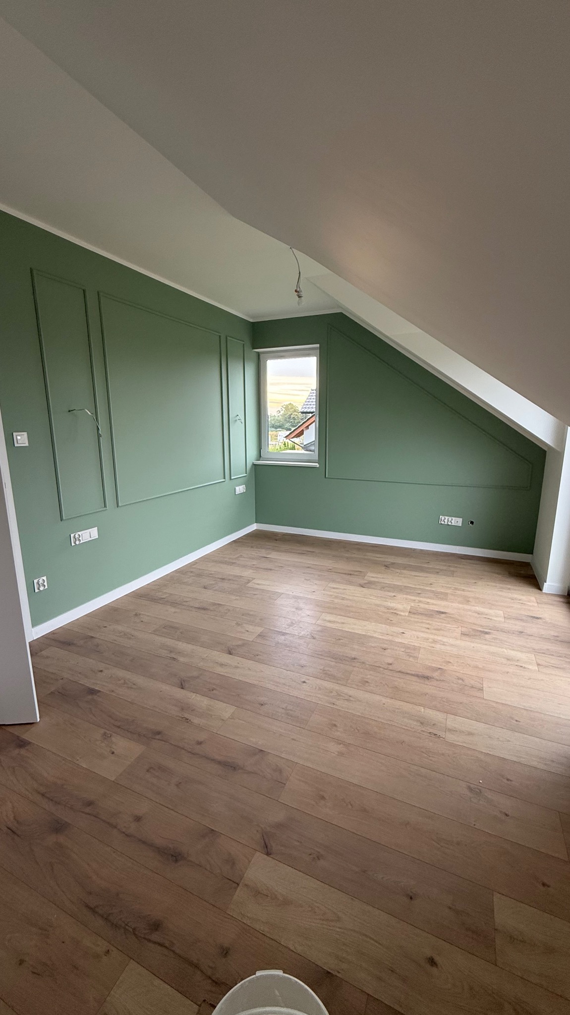 Pokój na poddaszu po remoncie: zielone ściany z dekoracyjnymi listwami, drewniana podłoga, okno z widokiem na zewnątrz. Skos dachu dodaje charakteru.