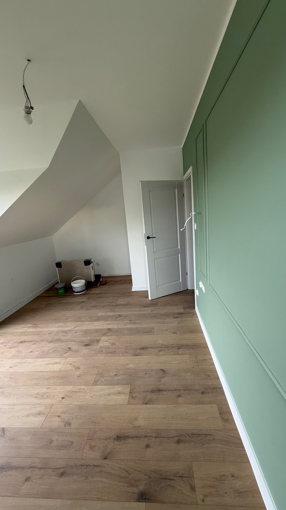 Pokój na poddaszu po remoncie: drewniana podłoga, zielona ściana z dekoracyjnymi listwami, białe ściany i drzwi. Widoczny sprzęt malarski.