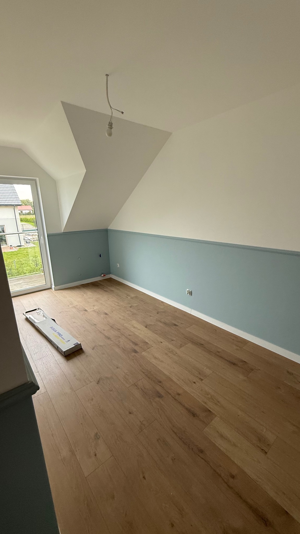 Pokój na poddaszu po remoncie: drewniana podłoga, pomalowane ściany na biało i błękitno, okno balkonowe. Na podłodze leży opakowanie paneli.