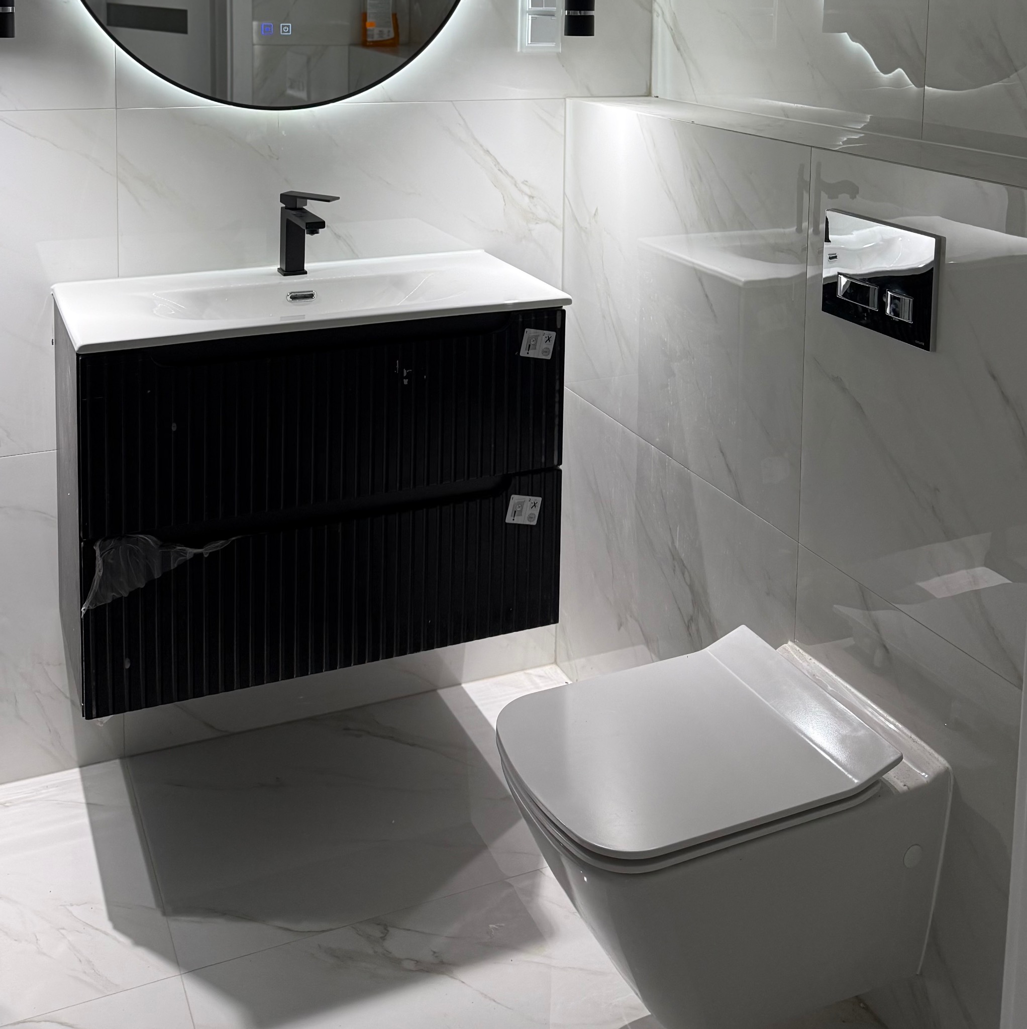 Nowoczesna łazienka z umywalką na szafce z czarnymi lamelami, podświetlanym lustrem i podwieszaną toaletą. Białe płytki imitujące marmur na ścianach i podłodze.