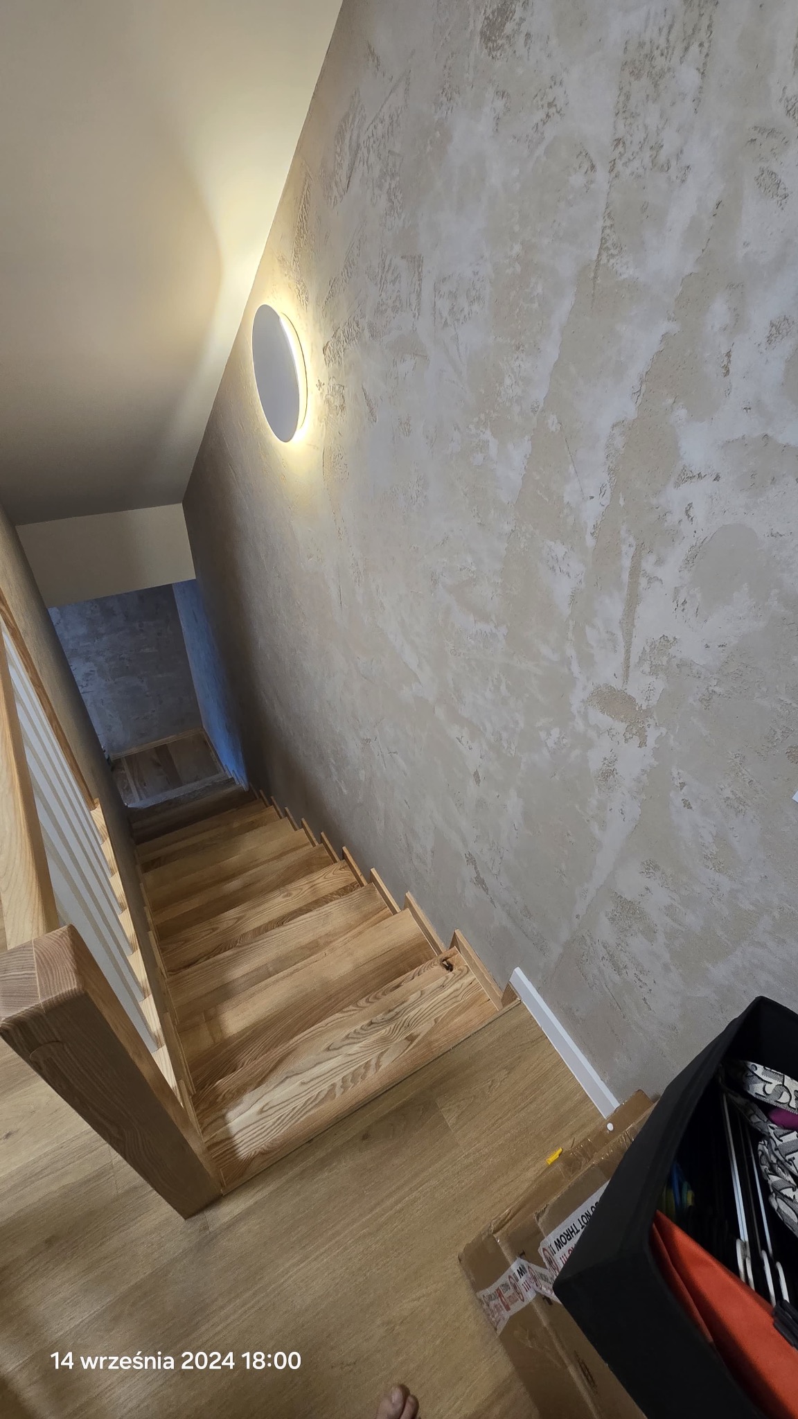 Drewniane schody z oświetleniem LED na ścianie o fakturze dekoracyjnej. Widok z góry, podkreślający naturalne materiały i nowoczesny design. Częściowo widoczny karton na podłodze.