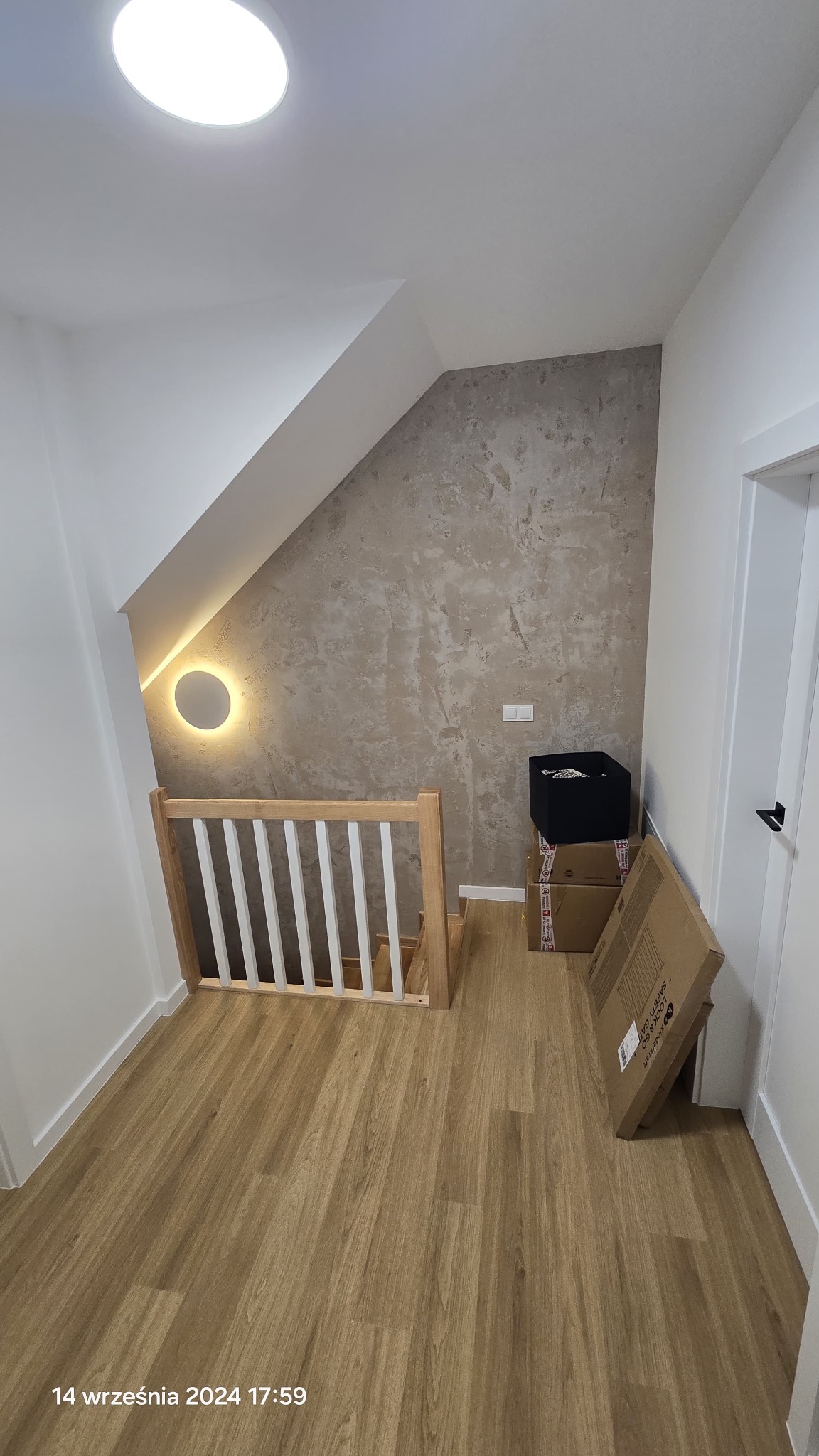 Wykończone poddasze z drewnianą barierką schodów, panelami podłogowymi i dekoracyjną ścianą z oświetleniem. Widoczne kartony i pudełko, sugerujące etap po remoncie.