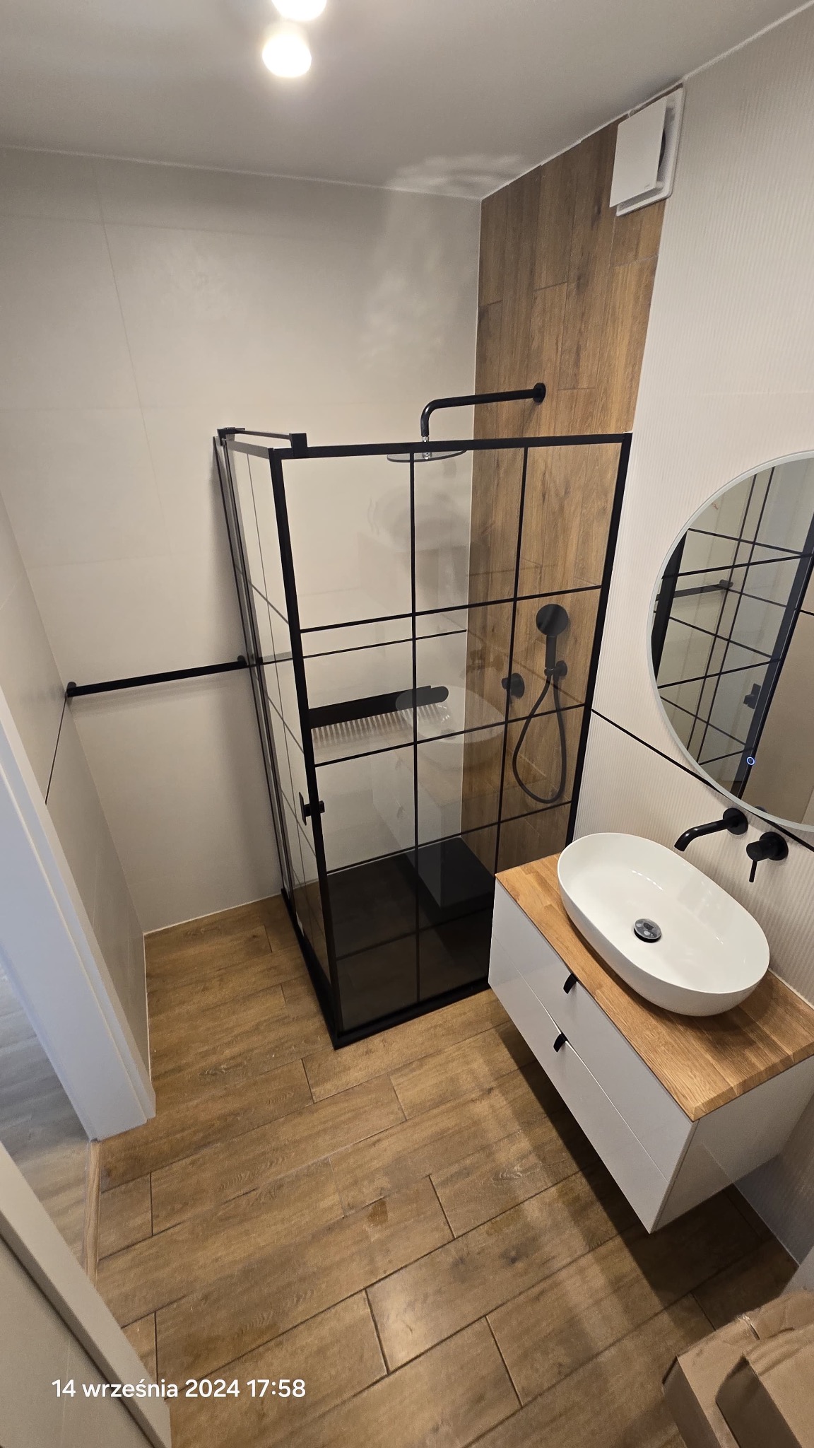 Nowoczesna łazienka po remoncie: kabina prysznicowa typu walk-in z czarnymi profilami, umywalka nablatowa na drewnianym blacie, lustro okrągłe z czarną ramą.