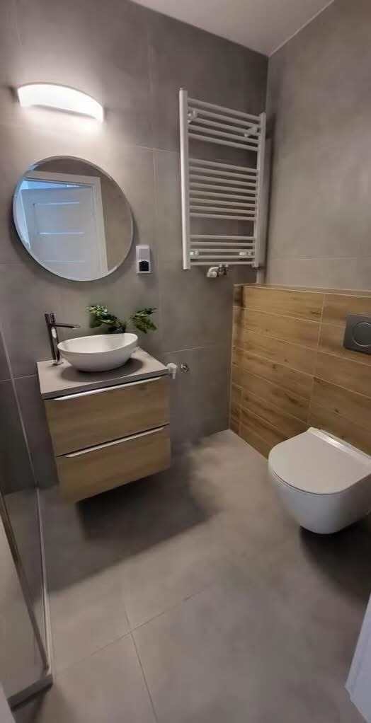 Nowoczesna łazienka z okrągłym lustrem, umywalką na szafce z drewnianymi frontami, grzejnikiem drabinkowym i płytkami imitującymi drewno na ścianie.