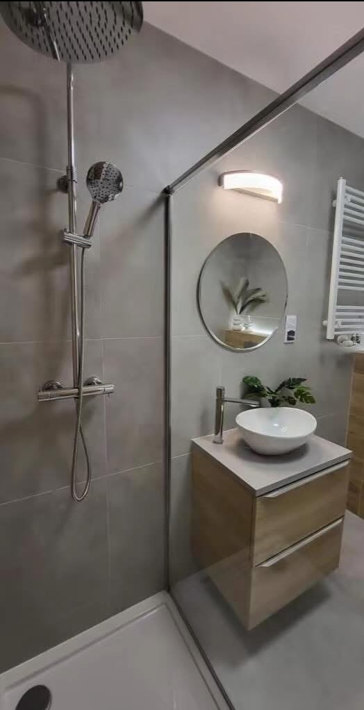 Nowoczesna łazienka z kabiną prysznicową typu walk-in, okrągłym lustrem i umywalką nablatową na szafce z drewnianymi frontami. Szare płytki na ścianach.