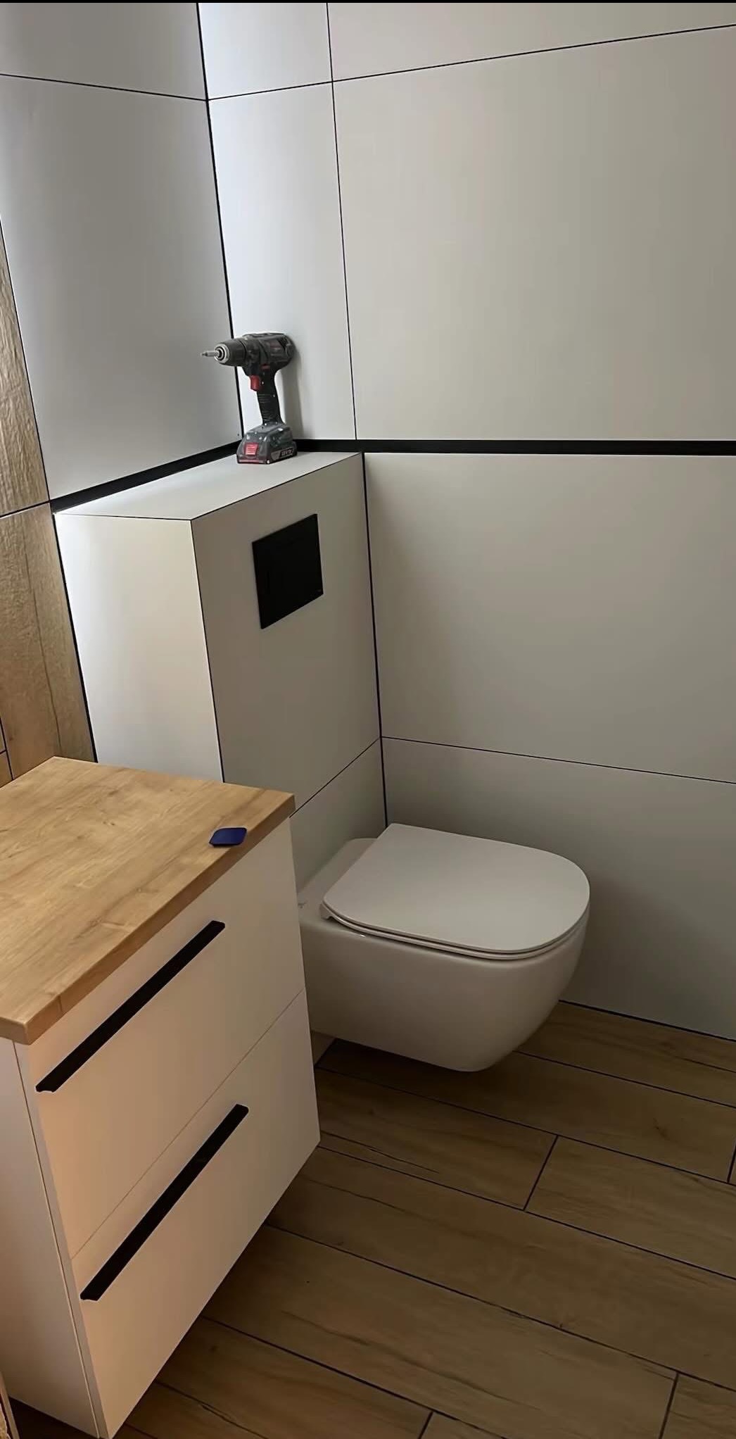 Nowoczesna łazienka po remoncie: białe płytki z czarnymi fugami, podwieszana toaleta, drewniana szafka z umywalką i wiertarka na obudowie. Minimalistyczny design.