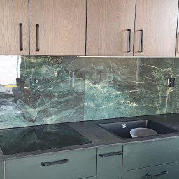 Dominik - Nowoczesna kuchnia z zielonymi szafkami i blatem, marmurowy panel nad blatem, czarne uchwyty i sprzęt AGD. Widok na fragment wnętrza z oknem.