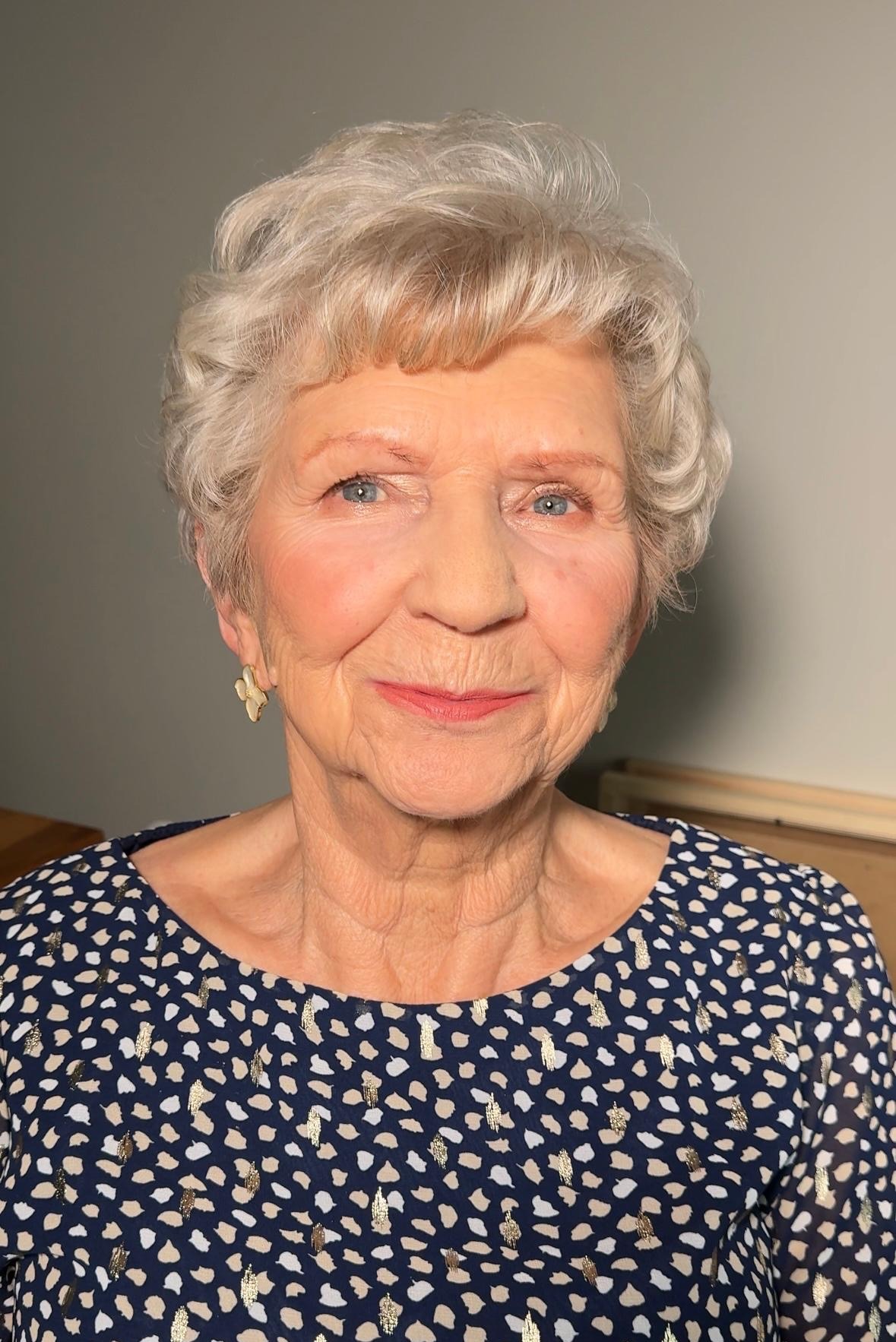 Portret starszej kobiety z delikatnym, okolicznościowym make-upem, podkreślającym rysy twarzy i kolor oczu. Widoczne kolczyki w kształcie kwiatów.