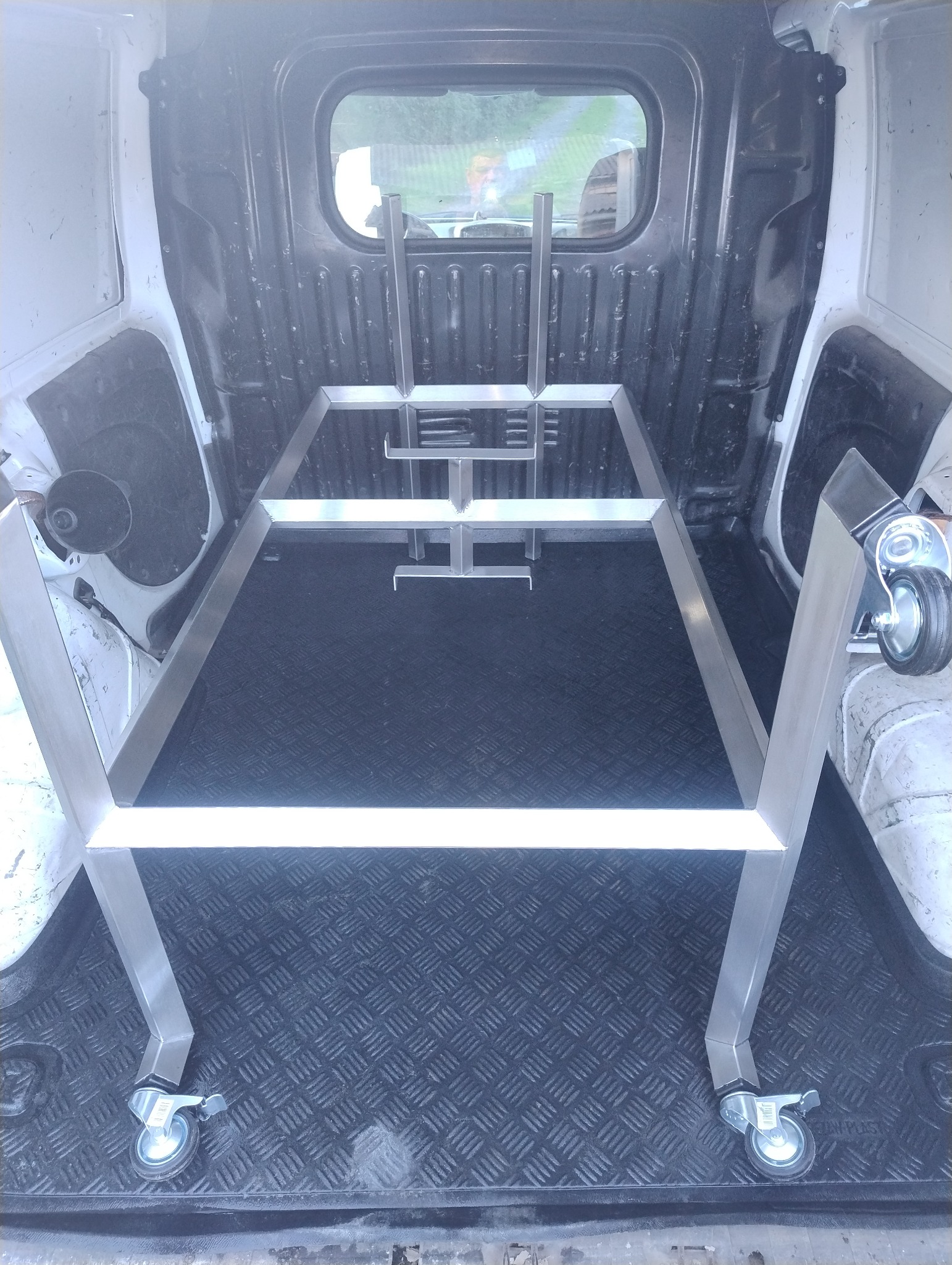 Metalowa konstrukcja na kółkach w przestrzeni ładunkowej samochodu dostawczego. Spawana rama z profili, widoczne spawy. Podłoga z gumową wykładziną.