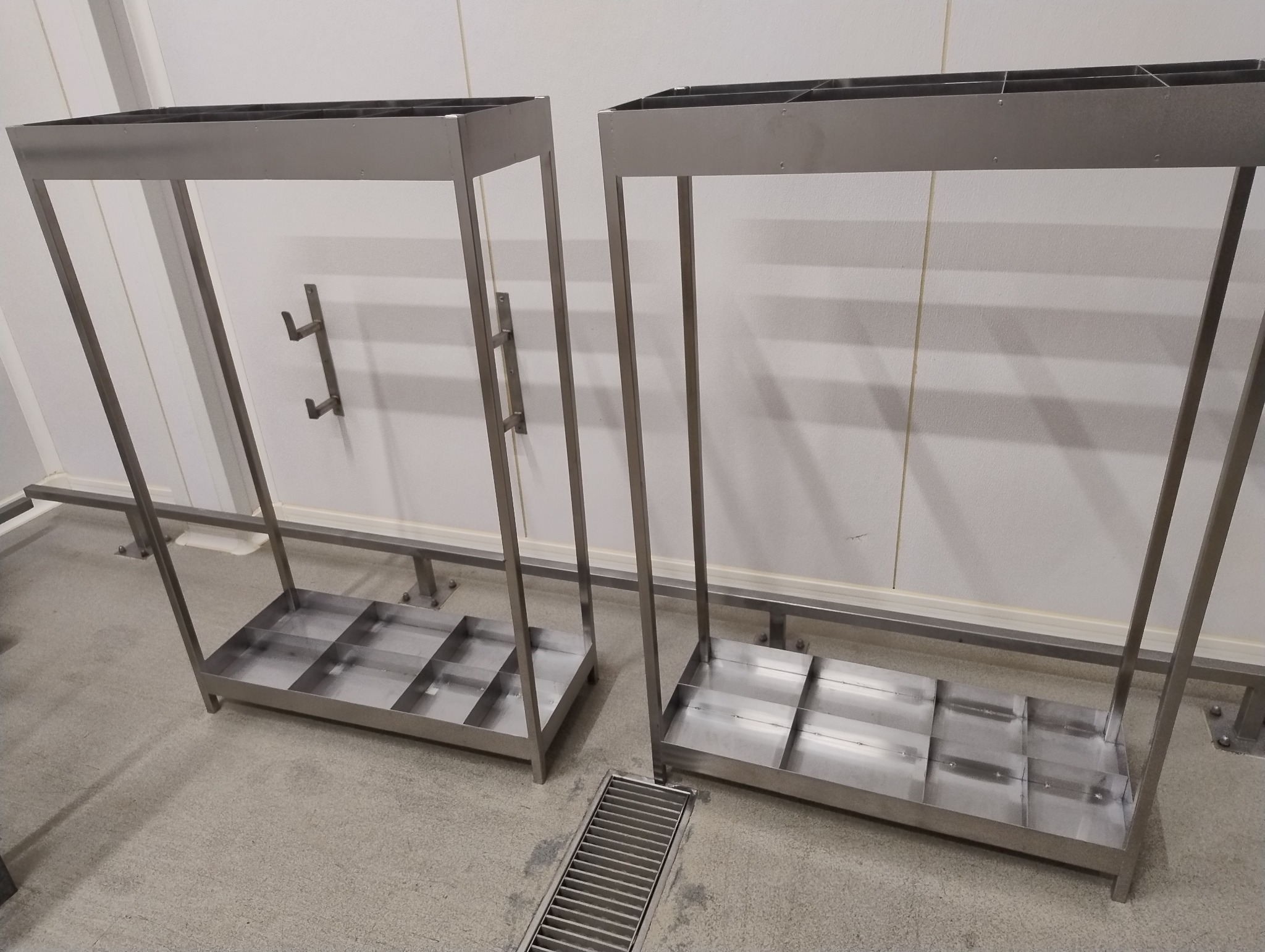 Dwa metalowe stojaki na parasole, wykonane ze stali nierdzewnej, z podziałem na przegródki, ustawione na szarym podłożu w jasnym pomieszczeniu.