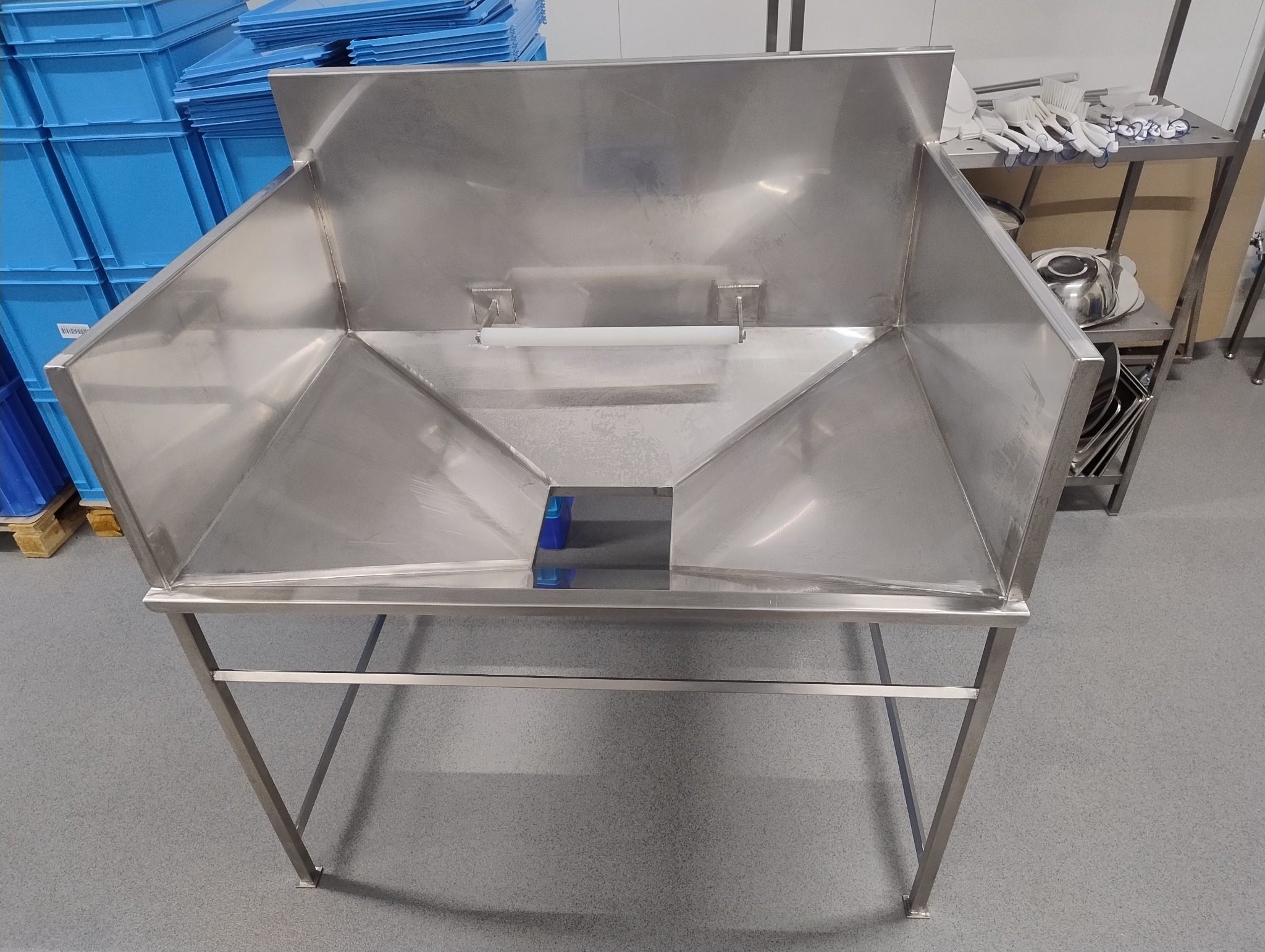 Stalowa konstrukcja na nogach, prawdopodobnie stół roboczy lub element wyposażenia przemysłowego, z widocznymi spawami i elementami montażowymi. W tle niebieskie pojemniki.