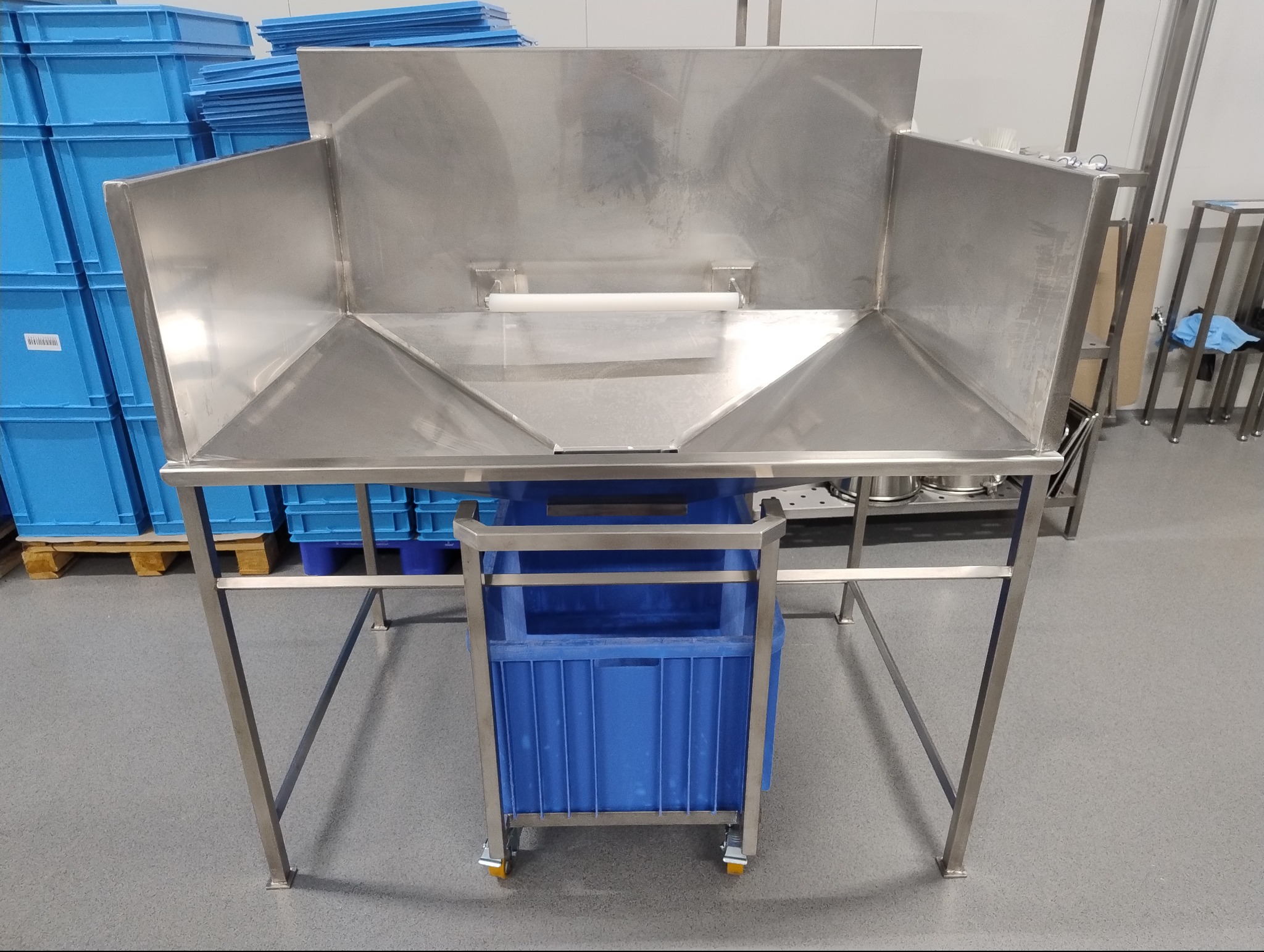 Stalowa konstrukcja stołu roboczego z wbudowanym zlewem i pojemnikiem na odpady, umieszczona w jasnym, przemysłowym otoczeniu z niebieskimi pojemnikami w tle.