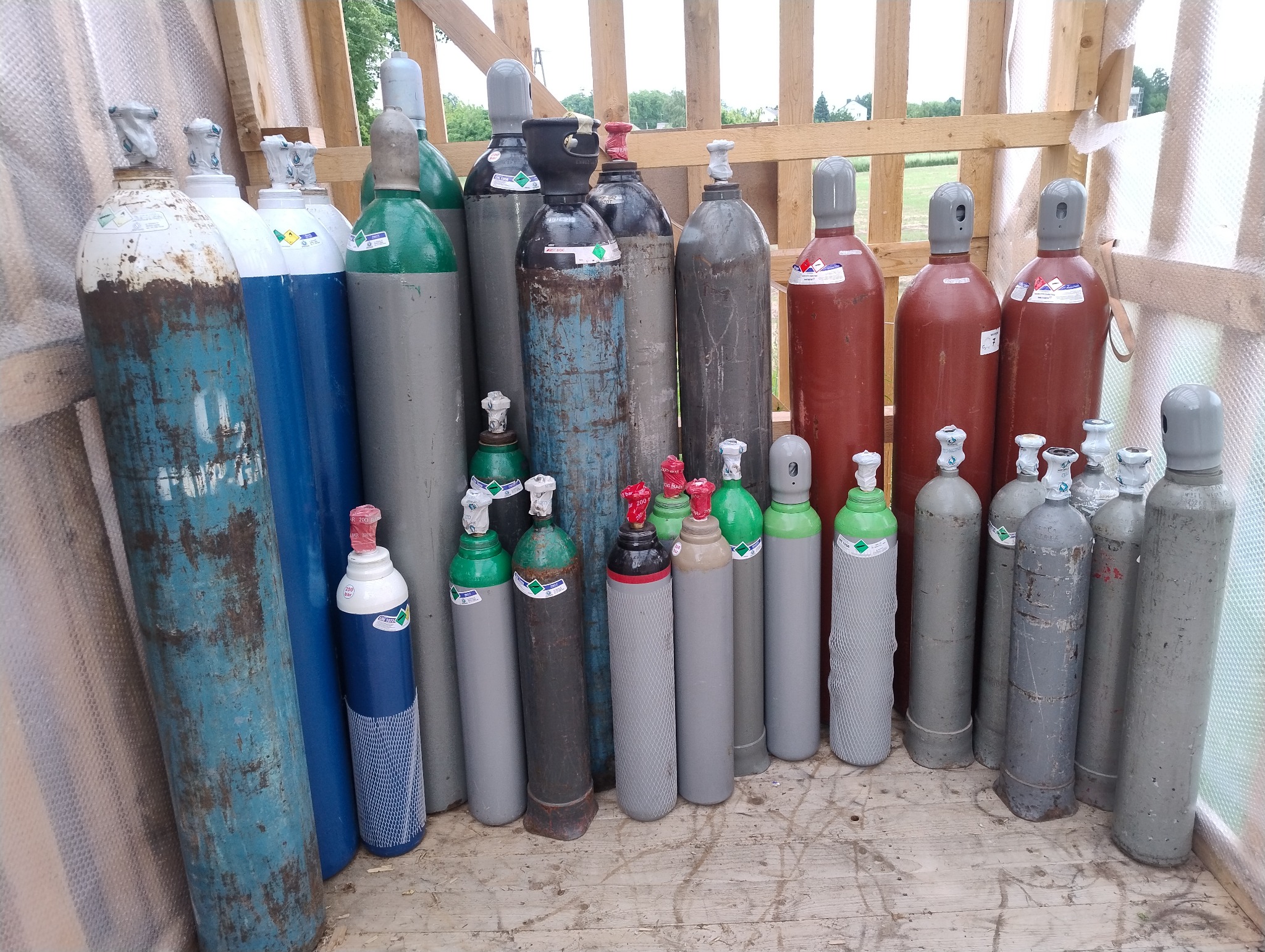 Różnokolorowe butle z gazem o różnych rozmiarach, ustawione w rzędach na drewnianej podłodze, niektóre z widocznymi etykietami i zabezpieczeniami.