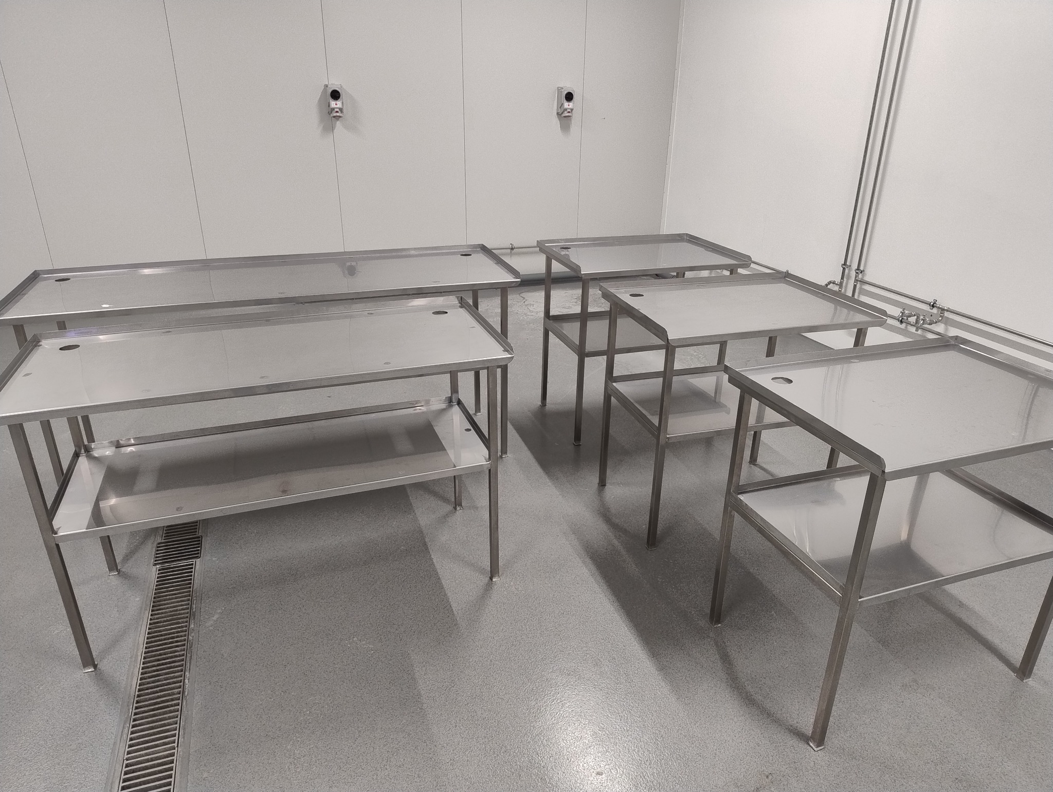 Metalowe stoły robocze ze stali nierdzewnej w sterylnym pomieszczeniu, widoczne rury i odpływ liniowy. Minimalistyczny design, równomierne oświetlenie.