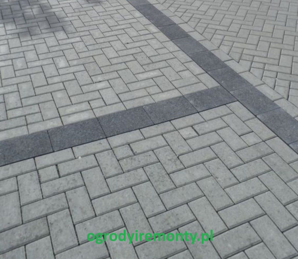 Ułożona szara kostka brukowa w jodełkę, obramowana ciemniejszym pasem kostki. Widok z góry, oświetlenie dzienne, widoczny fragment nawierzchni.