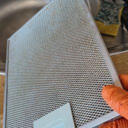 Sprzątanie A-Z - Filtr okapu kuchennego trzymany w dłoni w pomarańczowej rękawicy, widoczny zlew i gąbka. Filtr jest zabrudzony, co sugeruje potrzebę czyszczenia.