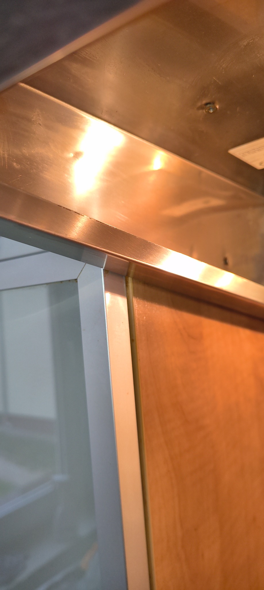 Detal wnętrza: narożnik szafki z aluminiową ramą i drewnianym frontem, oświetlony ciepłym światłem, widoczne ślady użytkowania na powierzchniach.