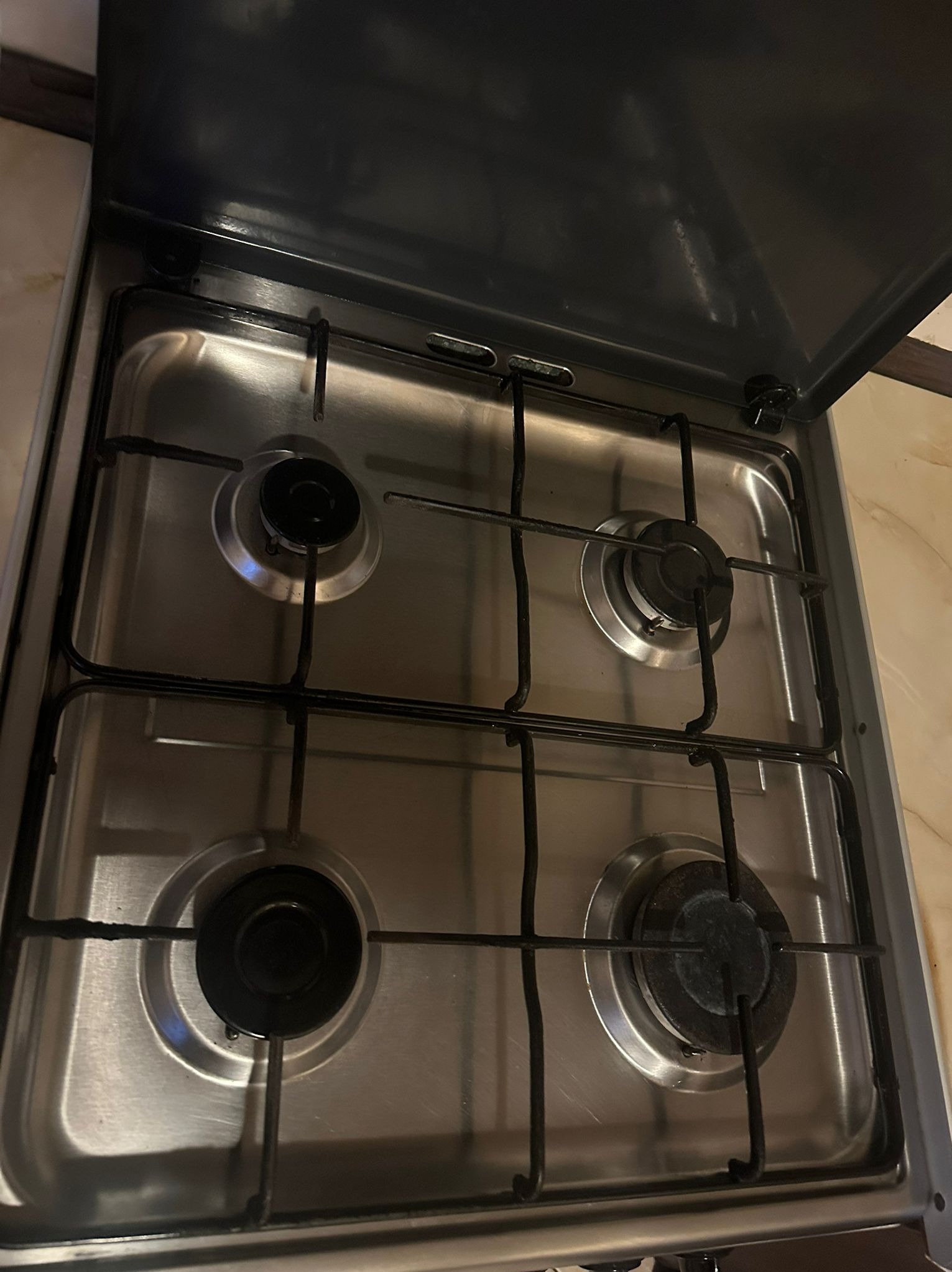 Czteropalnikowa, stalowa kuchenka gazowa z czarnymi rusztami i podniesioną pokrywą, widoczna z góry pod kątem. Palniki lekko zabrudzone, co sugeruje niedawne użycie.