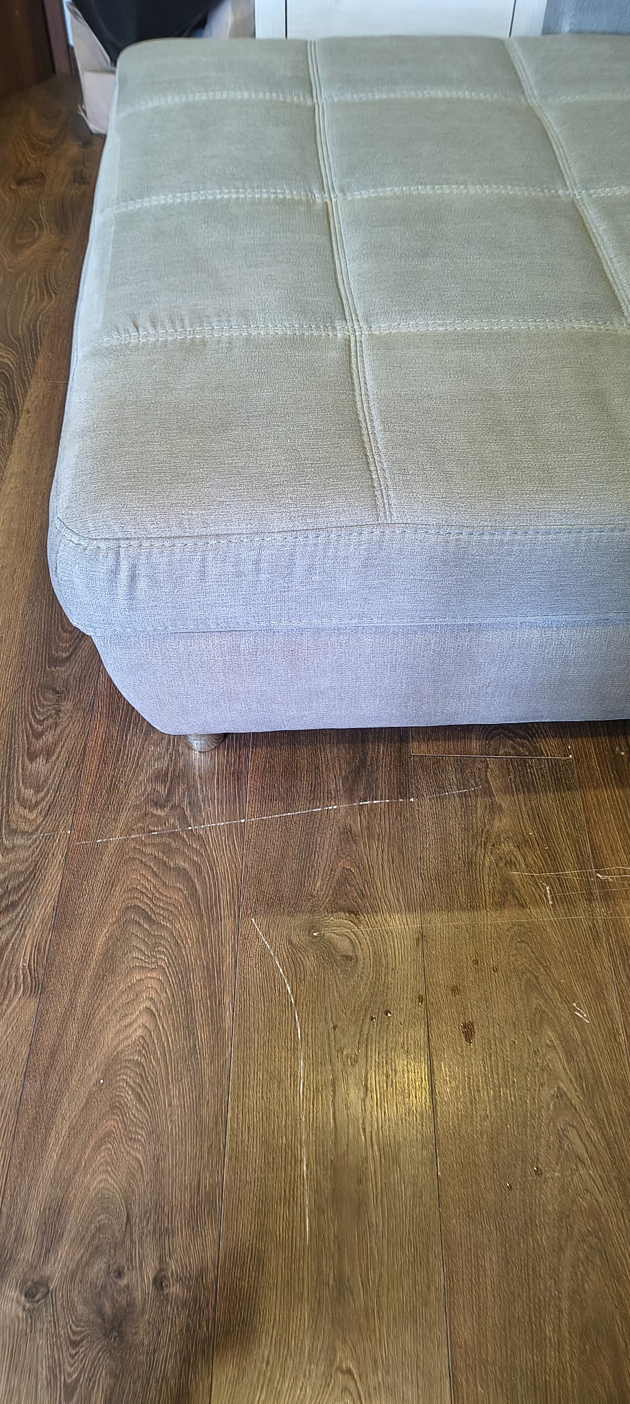 Jasna, tapicerowana pufa z widocznymi przeszyciami, stojąca na drewnianej podłodze z drobnymi zarysowaniami. Ujęcie z bliska, skupione na detalu tekstury i wykończenia.