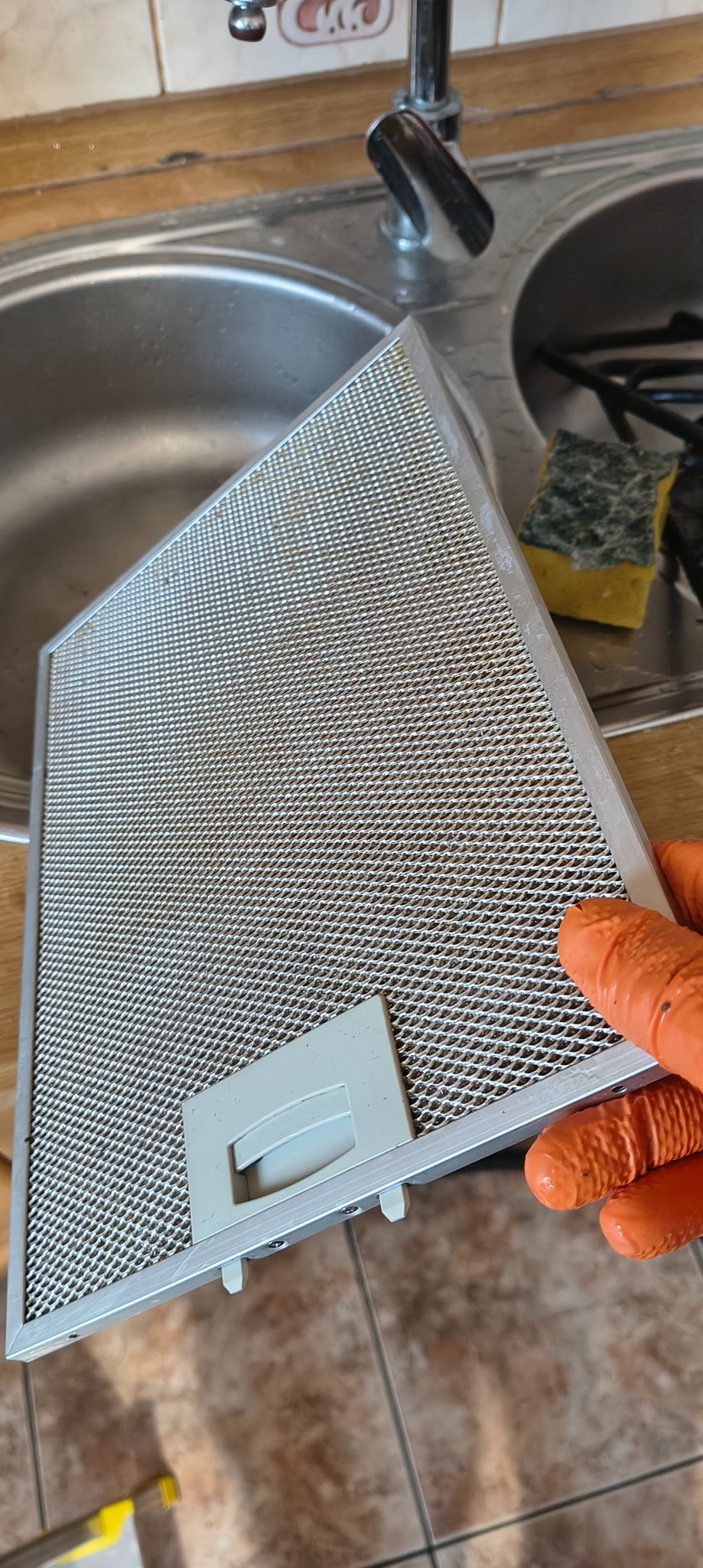 Filtr okapu kuchennego trzymany w dłoni w pomarańczowej rękawicy, widoczny zlew i gąbka. Filtr jest zabrudzony, co sugeruje potrzebę czyszczenia.