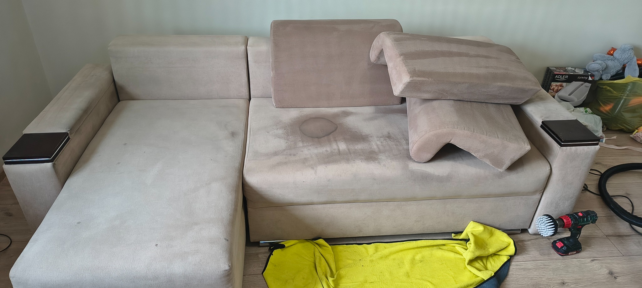 Beżowa, narożna kanapa z widocznymi plamami przed czyszczeniem. Na podłodze leży żółta ścierka i wiertarka z szczotką do czyszczenia tapicerki.