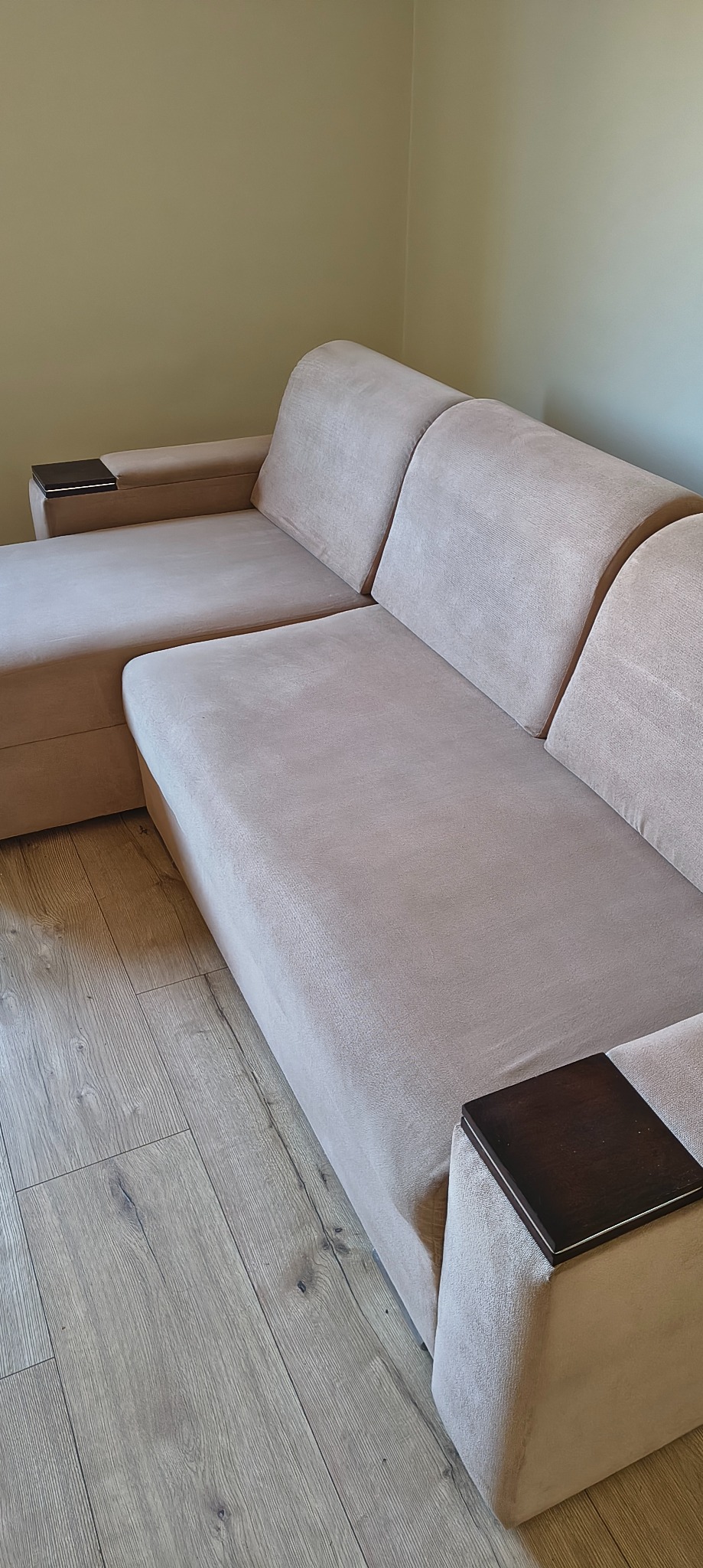 Beżowa, narożna kanapa z drewnianymi akcentami na podłogę z paneli. Widok z boku, prezentujący czyste obicie i nowoczesny design mebla.