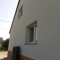 MatBud - Otynkowana elewacja domu z dwoma oknami, dolna część w kolorze brązowym. Obok ściany stoi klimatyzator na podeście i wiadro z farbą. Słoneczny dzień.