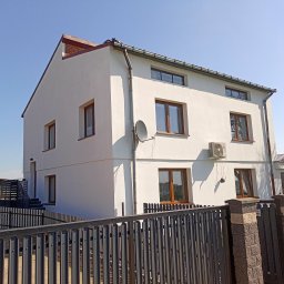 MatBud - Dwupiętrowy dom z białą elewacją i brązowymi oknami, widoczny zza metalowego ogrodzenia. Na ścianie antena satelitarna i klimatyzator. Błękitne niebo w tle.