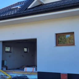 MatBud - Biała elewacja domu jednorodzinnego po tynkowaniu, z widoczną drabiną opartą o rynnę i otwartym garażem. Okno z drewnianą ramą odbija krajobraz.