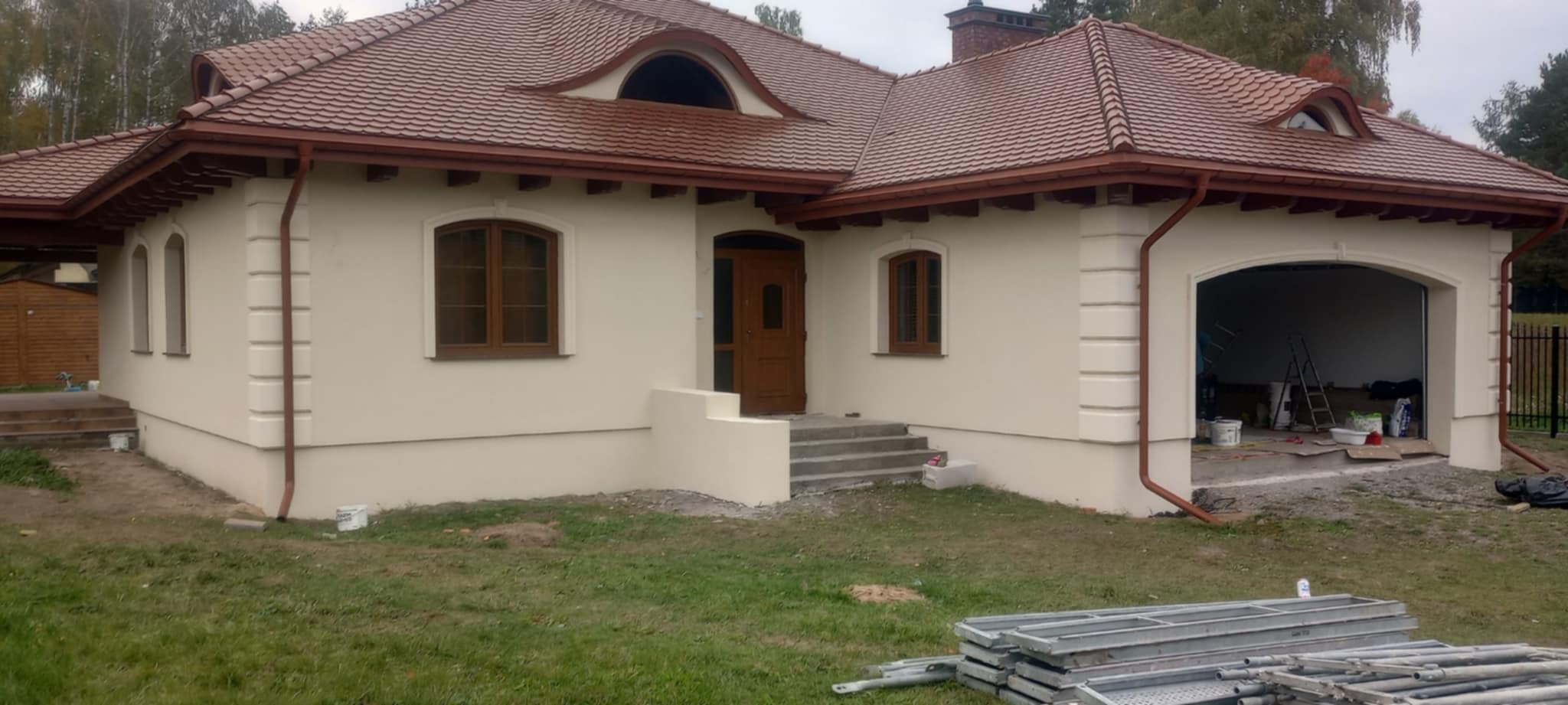 Elewacja domu jednorodzinnego w jasnym odcieniu z brązową stolarką okienną i dachem z dachówki. Widoczne rusztowania i elementy wykończeniowe.