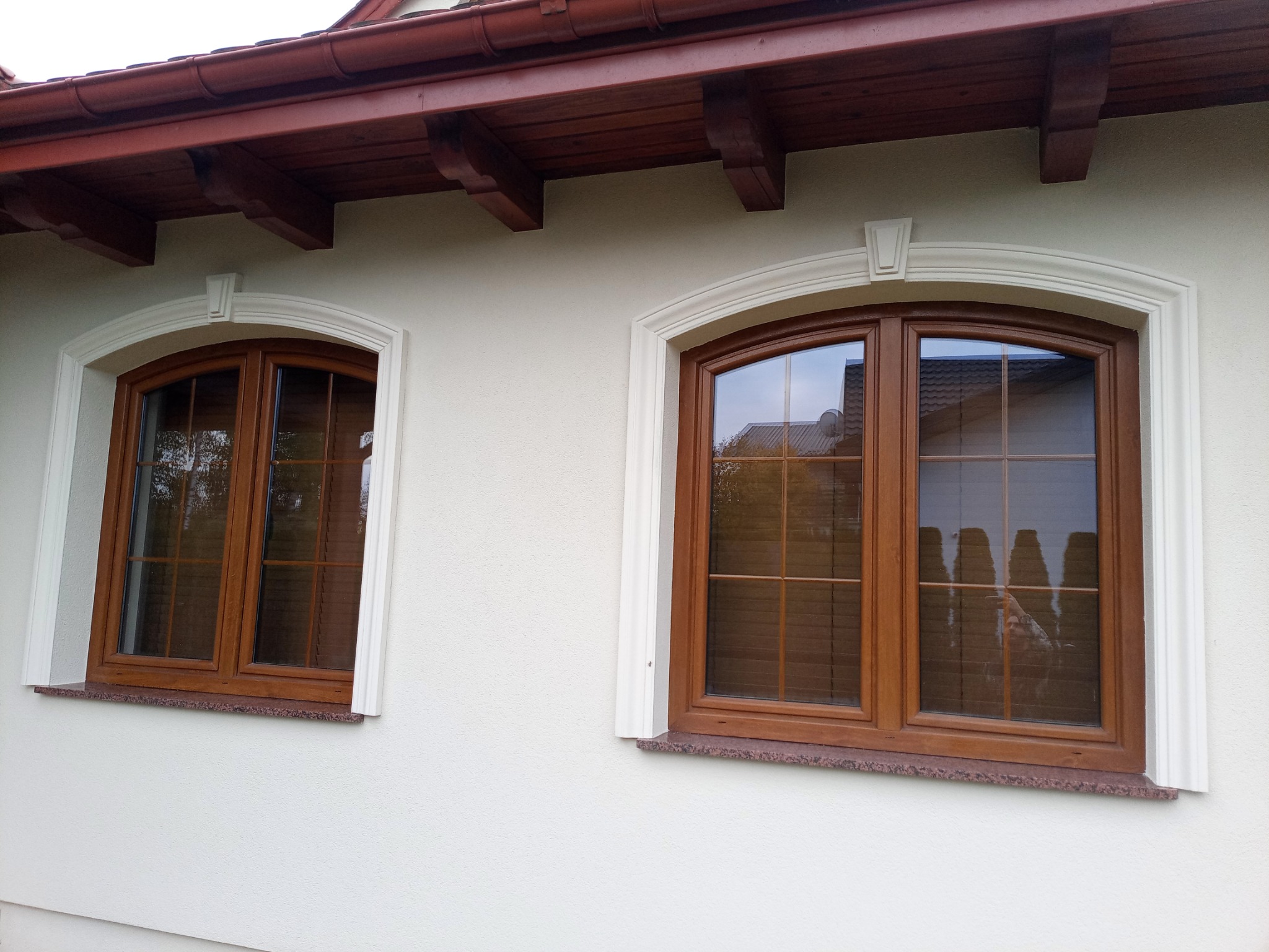 Dwa okna z brązowymi ramami i białymi ozdobnymi obramowaniami na elewacji budynku. Widoczny fragment dachu z brązowymi rynnami.