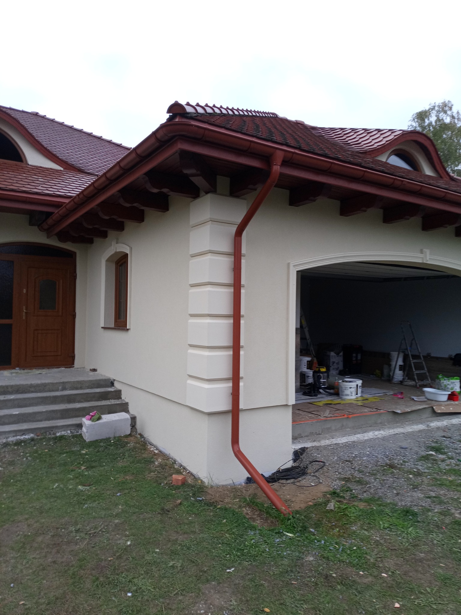 Wykończona elewacja domu jednorodzinnego z brązową stolarką okienną i drzwiami. Widoczny fragment dachu z brązową dachówką i rynną. W tle otwarty garaż z narzędziami.