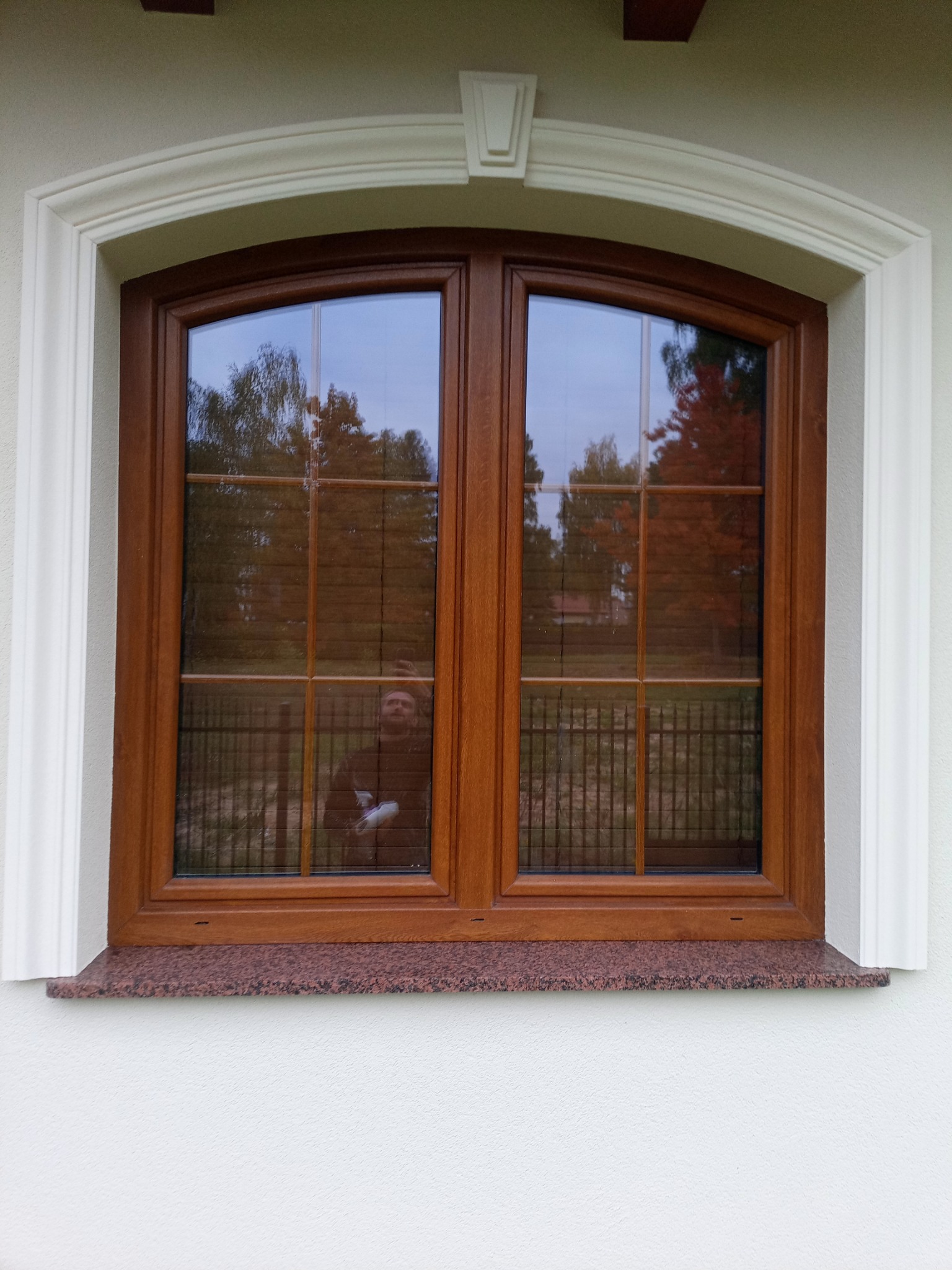 Drewniane okno z ozdobną, białą ramą i parapetem z czerwonego granitu, odbicie osoby w szybie. Elewacja budynku w jasnym kolorze.