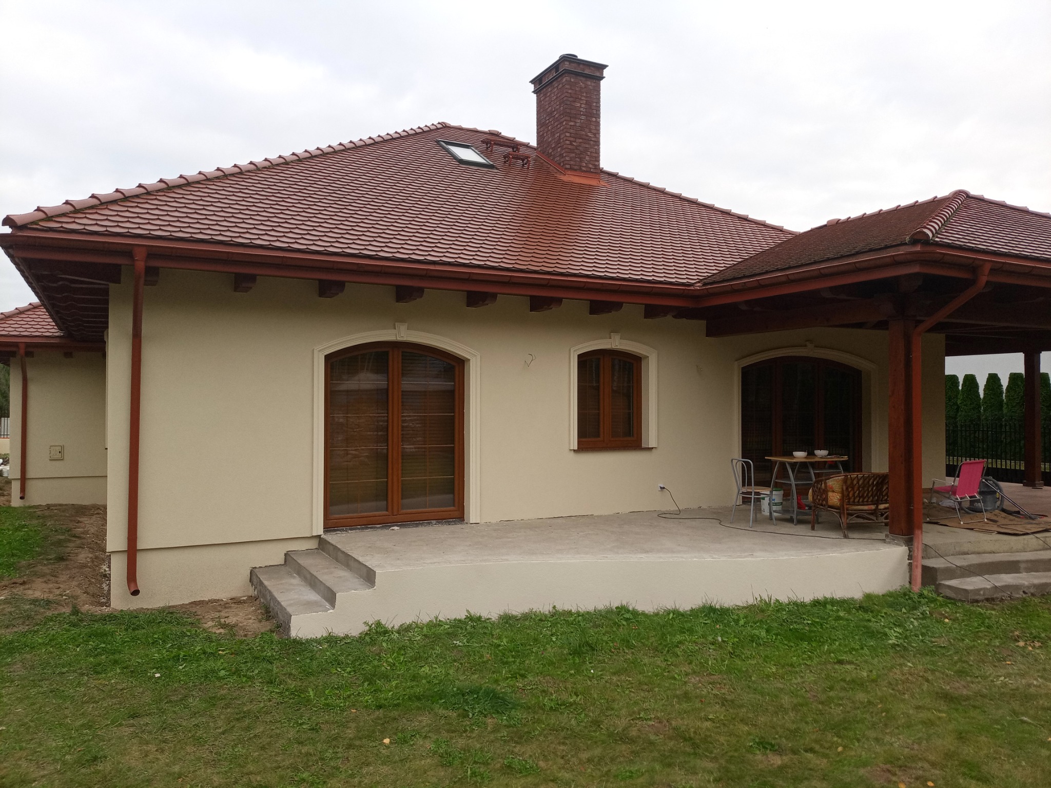 Dom jednorodzinny z beżową elewacją, czerwoną dachówką i ceglanym kominem. Widoczne okna z brązowymi ramami i fragment tarasu z meblami ogrodowymi.