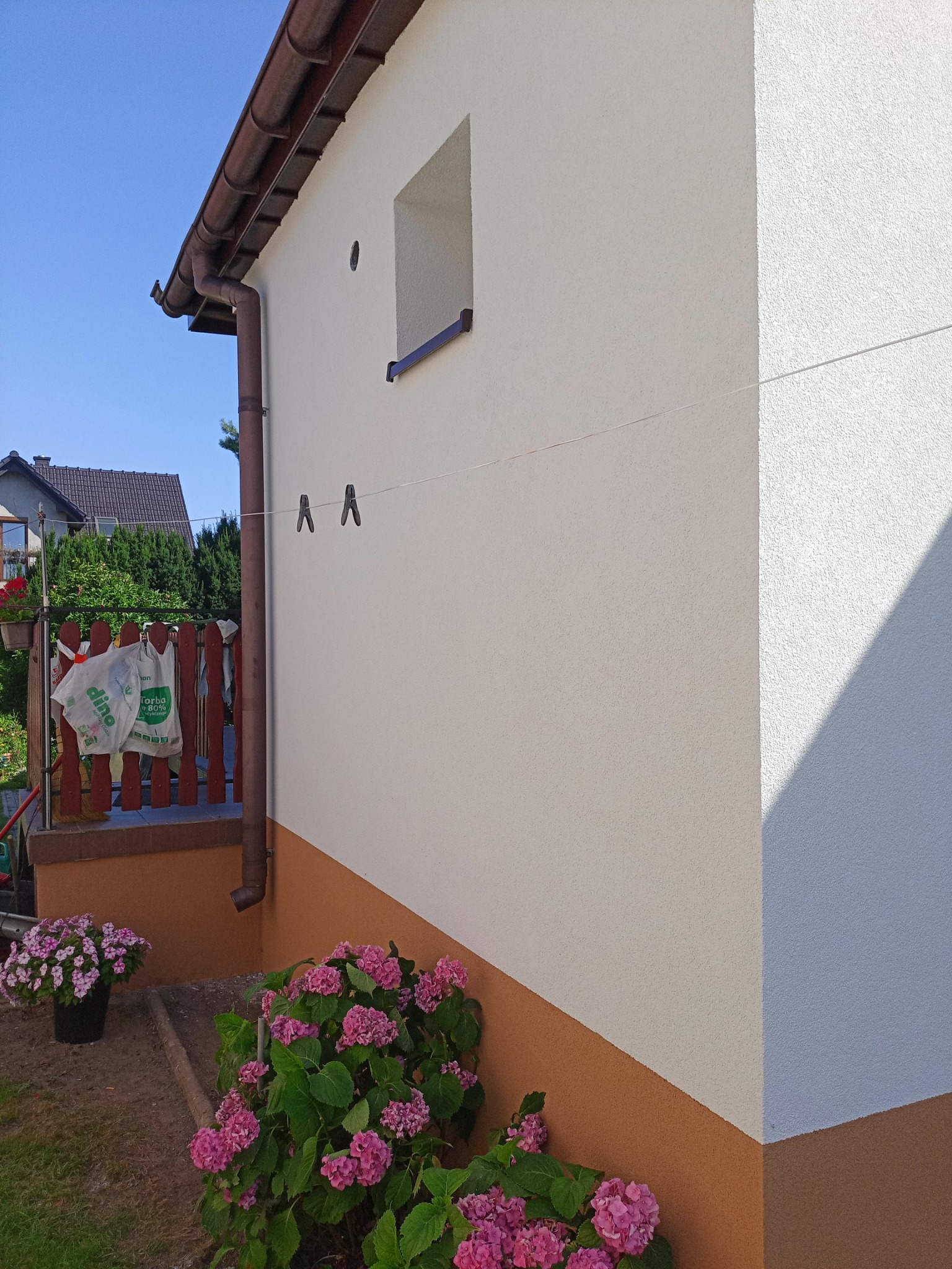 Ściana budynku po tynkowaniu elewacji, z widocznym fragmentem rynny, oknem i kwiatami. Dół ściany w kolorze brązowym, góra w odcieniach bieli. Dwa klamerki na sznurku.