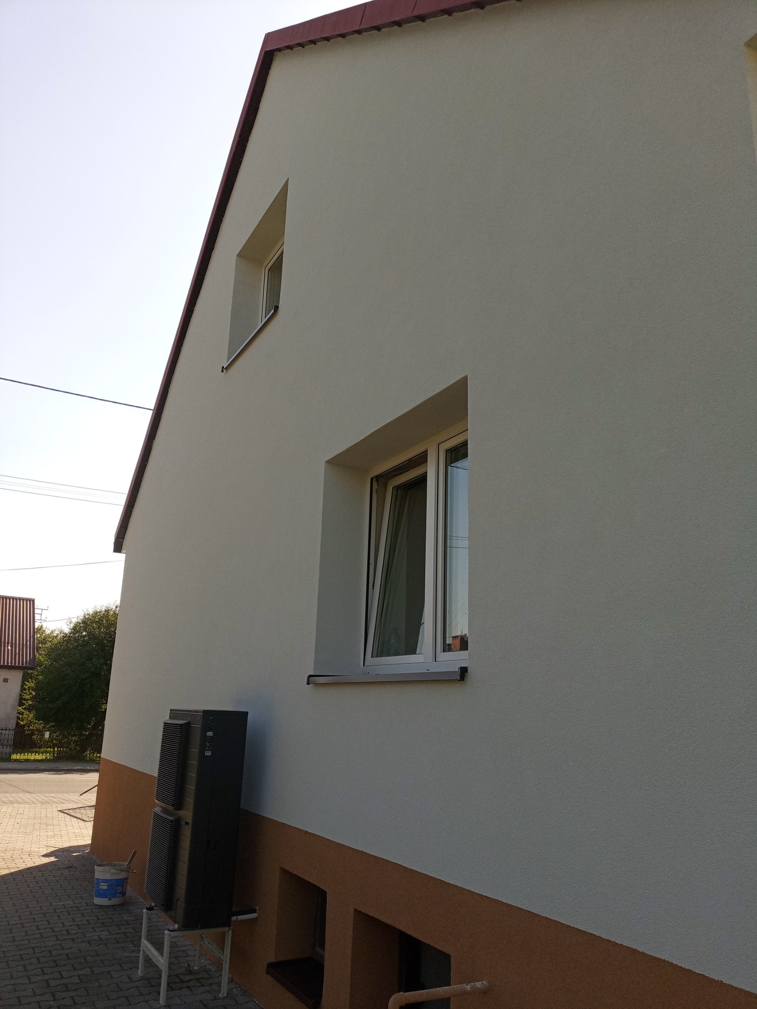 Otynkowana elewacja domu z dwoma oknami, dolna część w kolorze brązowym. Obok ściany stoi klimatyzator na podeście i wiadro z farbą. Słoneczny dzień.