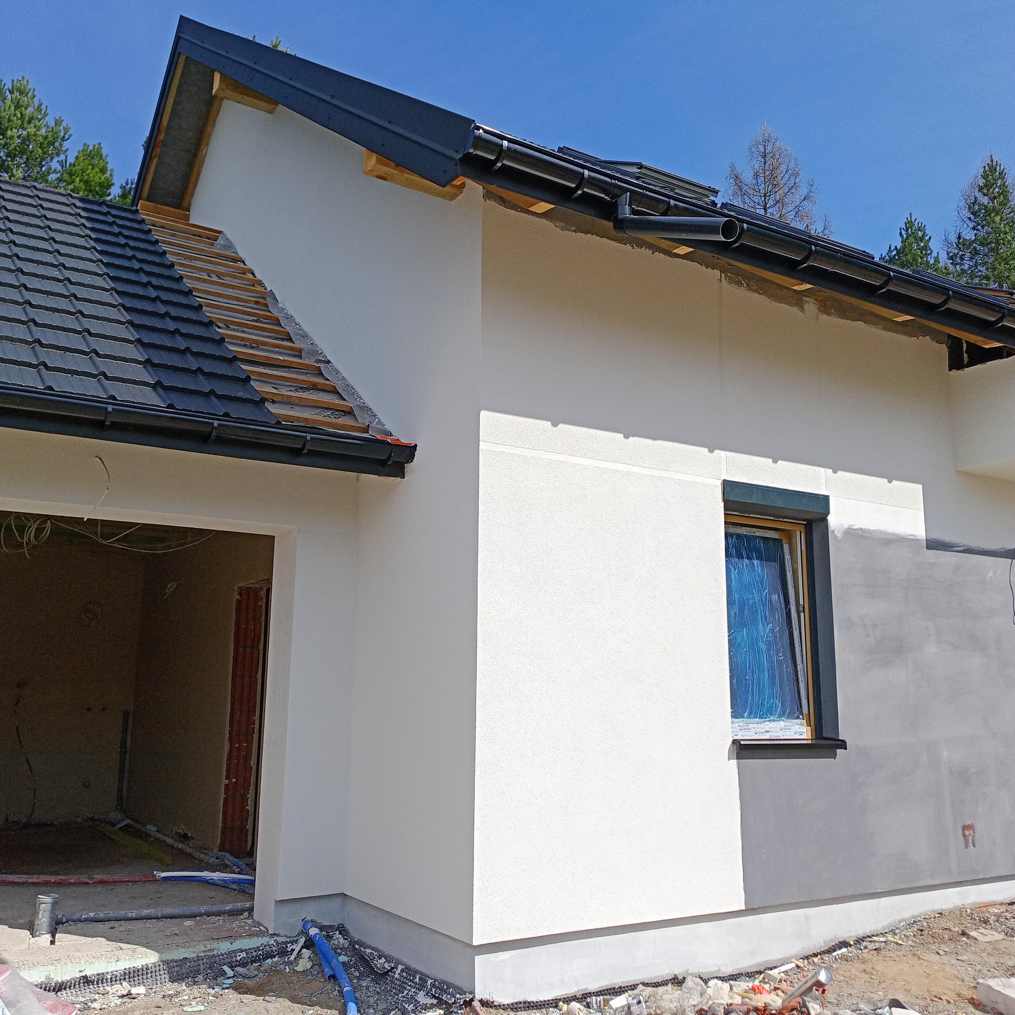 Fragment elewacji domu jednorodzinnego w trakcie prac wykończeniowych. Widoczna biała, otynkowana ściana, okno zabezpieczone folią i fragment dachu z czarną dachówką.