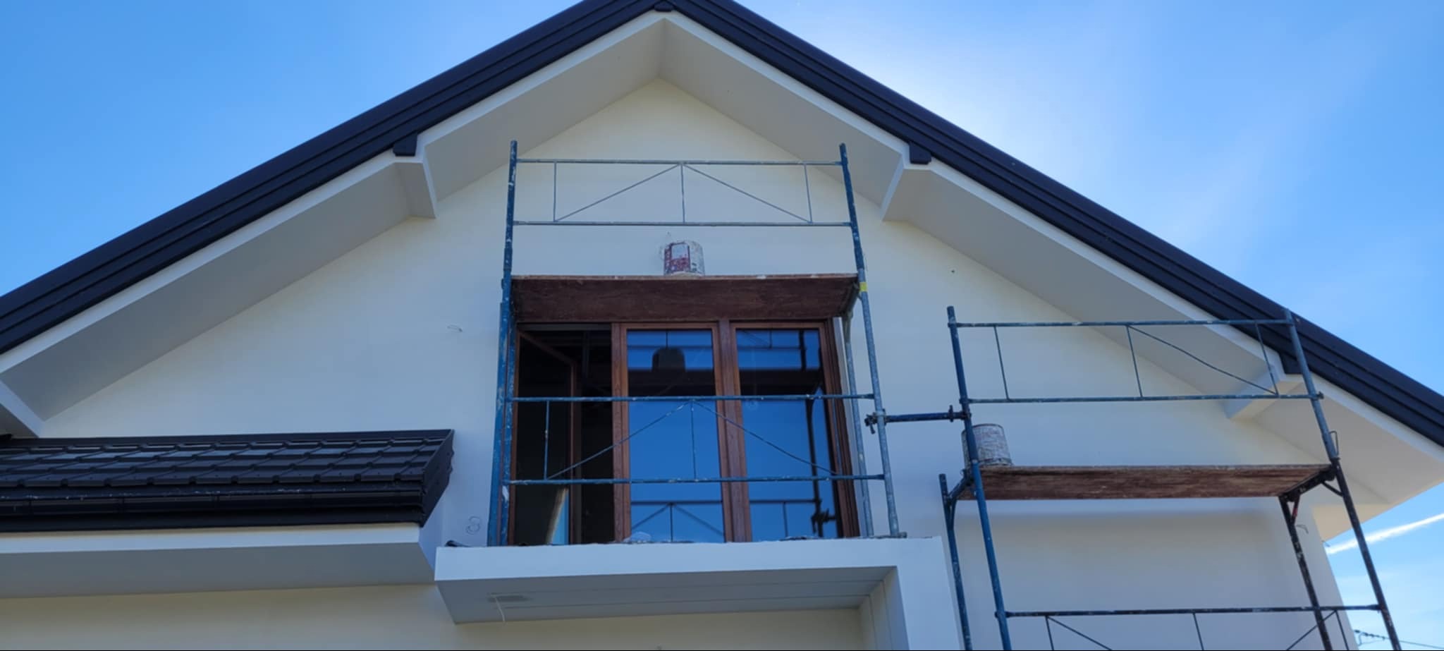 Biała elewacja domu jednorodzinnego w trakcie prac wykończeniowych, widoczne rusztowania i okno balkonowe z drewnianą ramą. Błękitne niebo w tle.