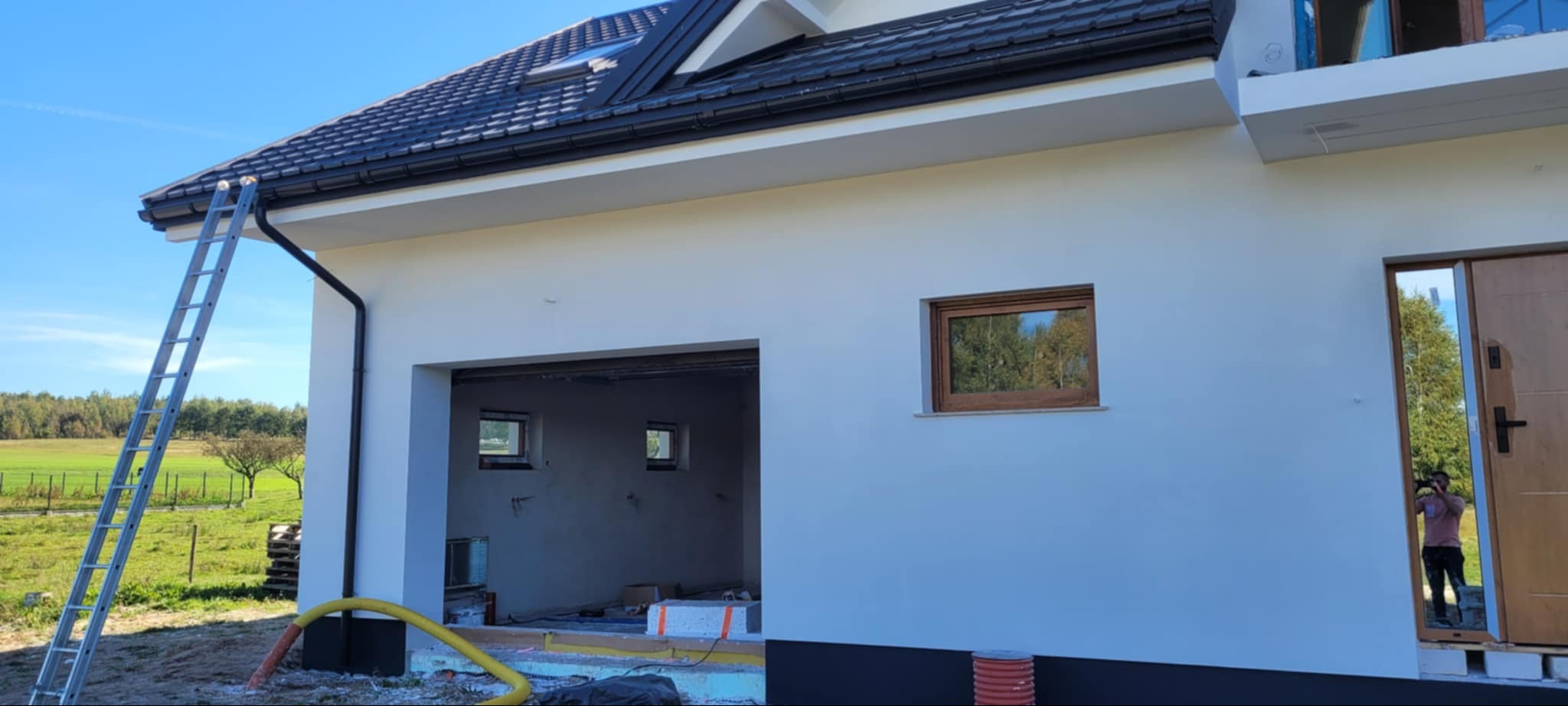 Biała elewacja domu jednorodzinnego po tynkowaniu, z widoczną drabiną opartą o rynnę i otwartym garażem. Okno z drewnianą ramą odbija krajobraz.