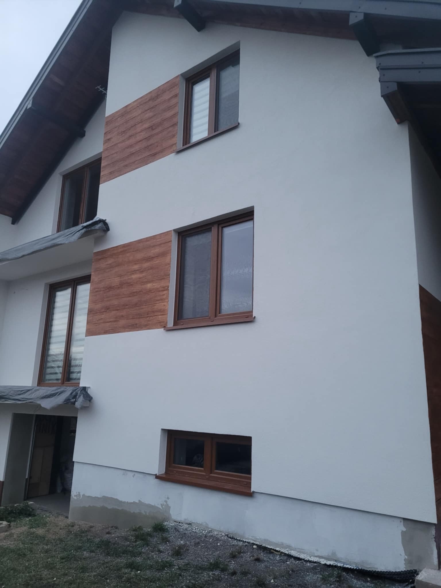 Wykończona elewacja domu z białym tynkiem i dekoracyjnymi panelami imitującymi drewno. Widoczne brązowe ramy okienne i fragment dachu.