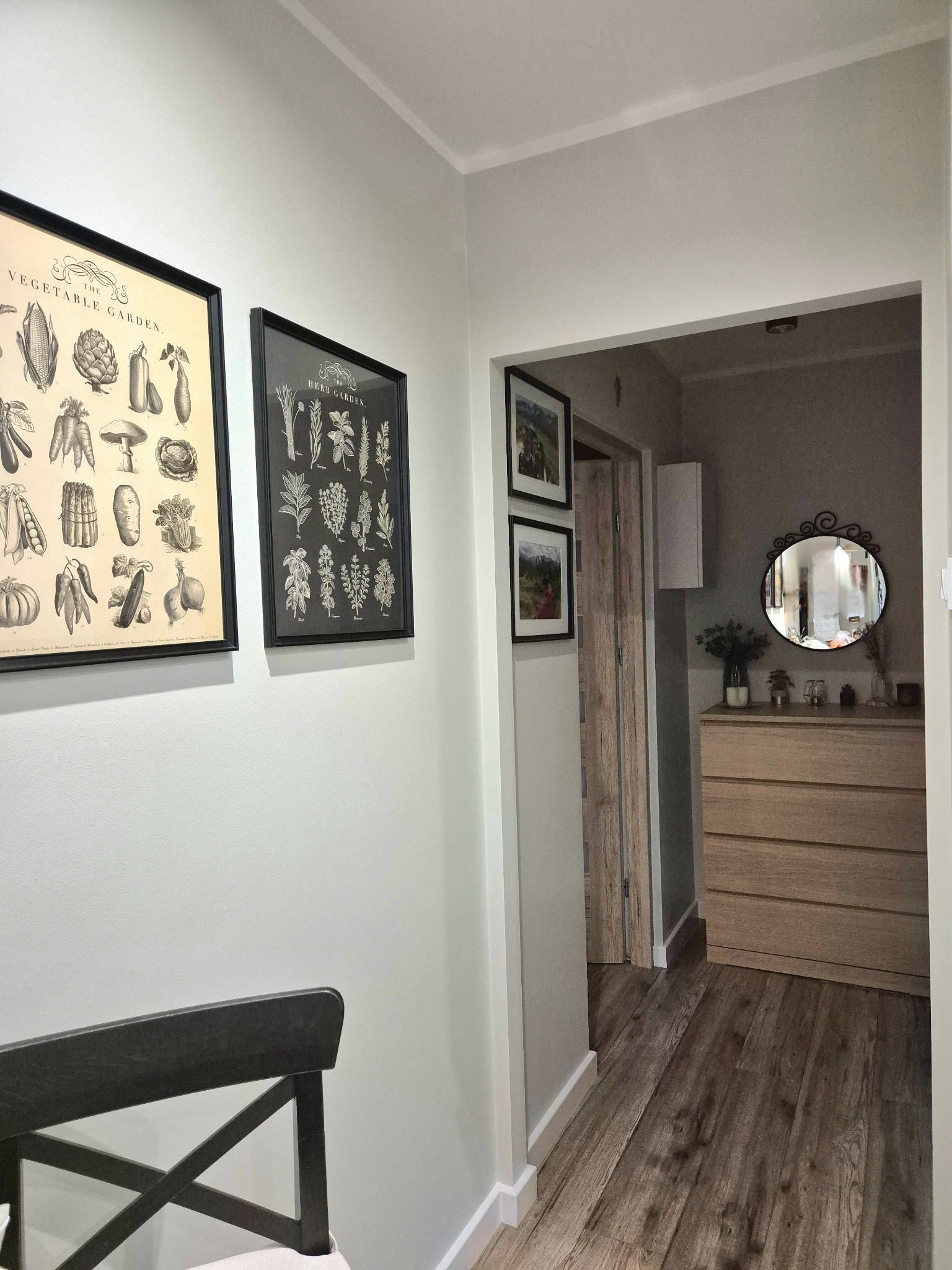 Jasny korytarz z drewnianą podłogą, ozdobiony grafikami w czarnych ramkach i komodą z lustrem. Widoczne detale wykończenia wnętrz.