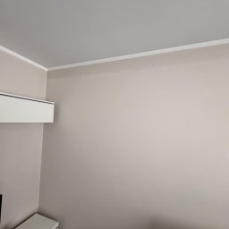 Krystian Sudoł - Ściana w odcieniach beżu po malowaniu, z białą listwą przysufitową i klimatyzatorem. Minimalistyczne wnętrze z elementami mebli w dolnej części kadru.