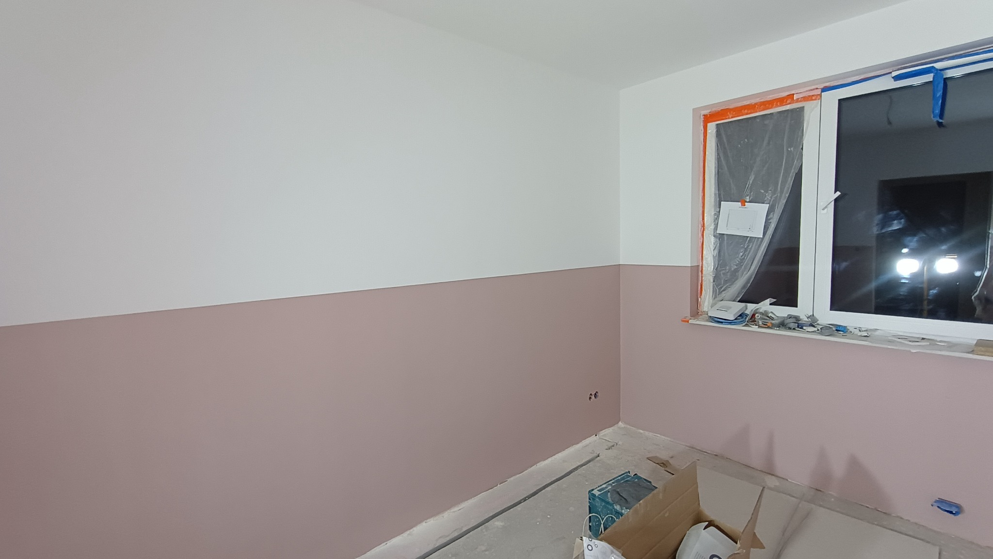 Pomieszczenie po malowaniu ścian na dwa kolory: biały u góry i pudrowy róż na dole. Okno zabezpieczone folią malarską. Widoczne puszki i narzędzia malarskie.